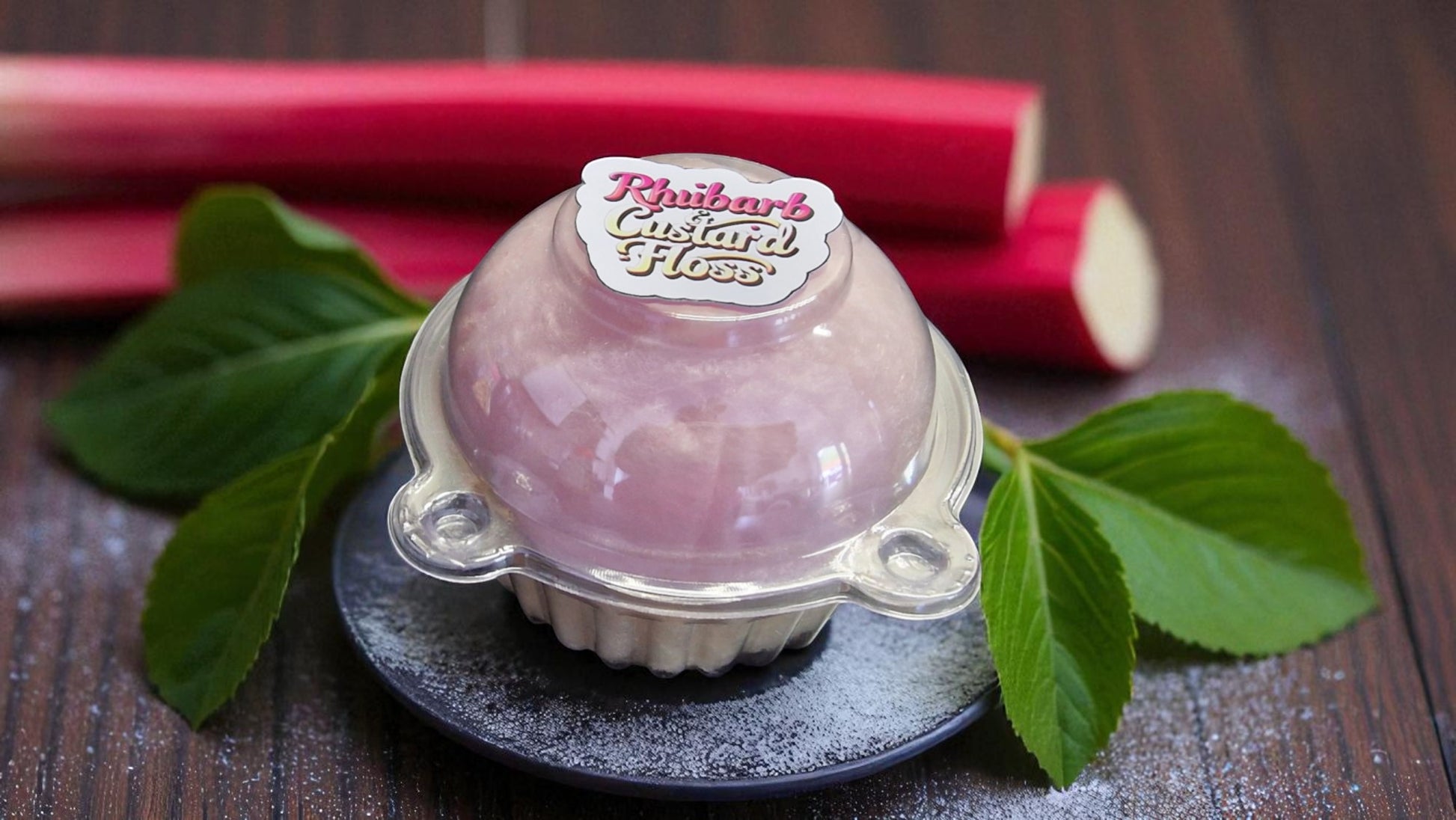 Pink custard flan in a clear dome packaging with rhubarb and leaves on a wooden surface