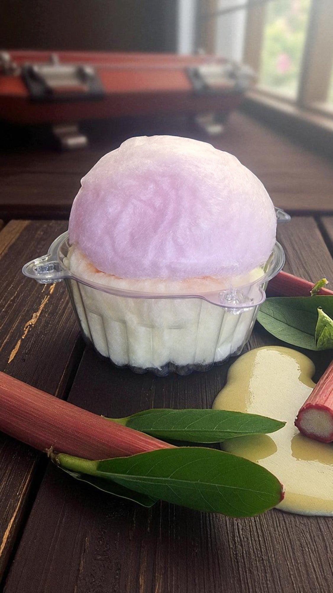 Pink ice cream scoop in a transparent container on a wooden surface with leaves and rhubarb.