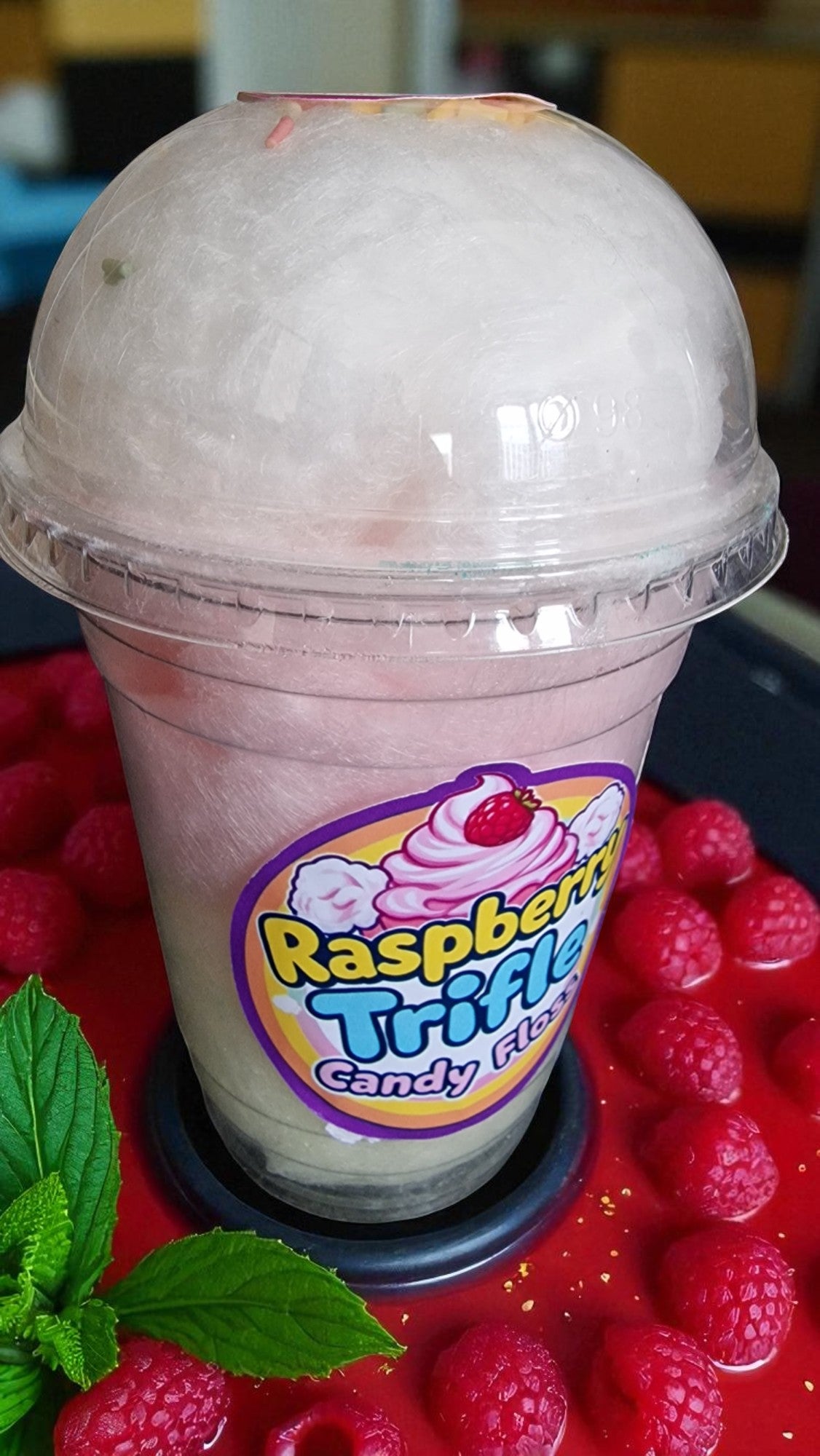 Pink drink with a Raspberry Trifle Candy Floss label on a bed of raspberries.