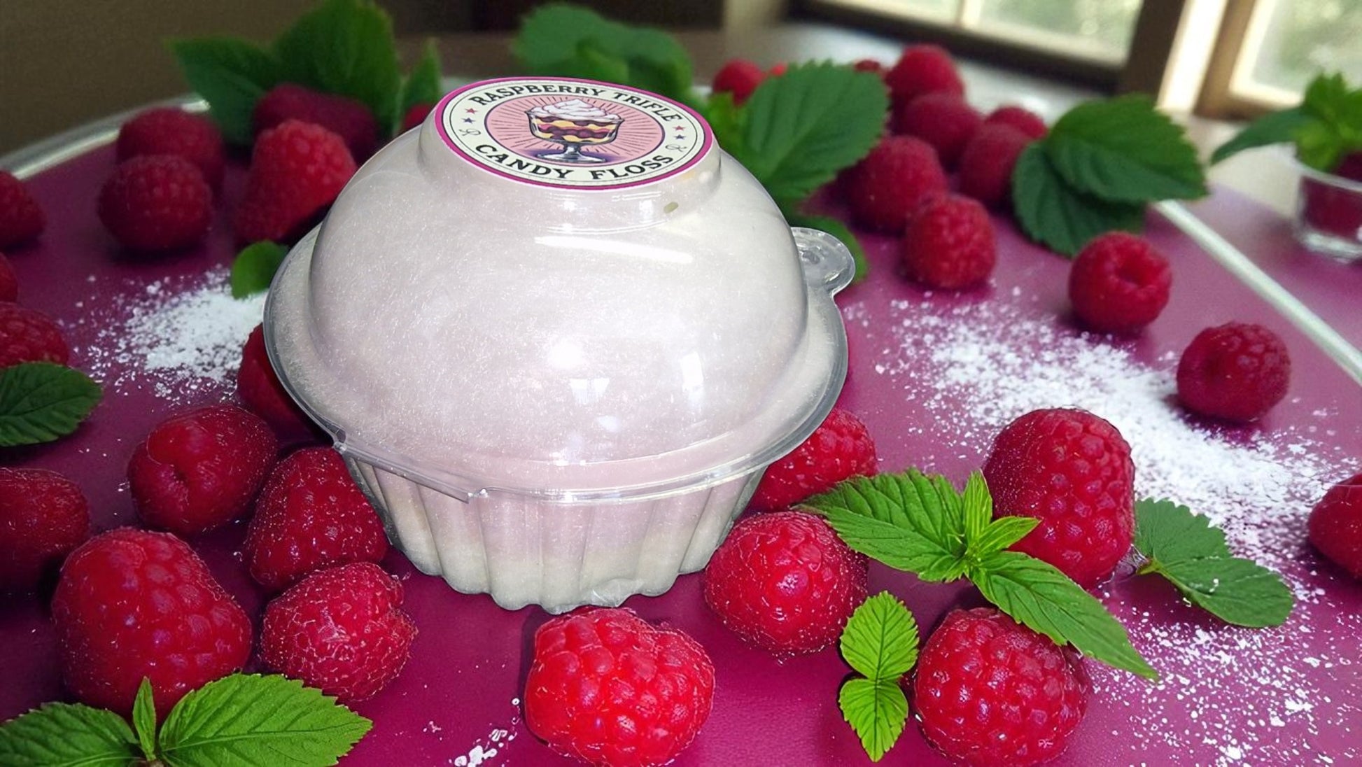 Raspberry flavored candy fluff in a container surrounded by fresh raspberries on a pink surface.
