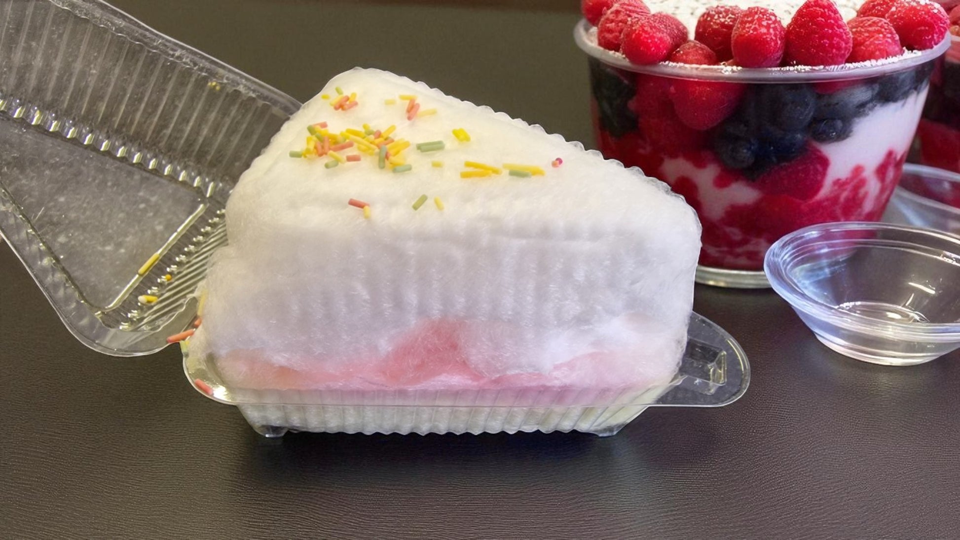 White dessert with colorful sprinkles in a clear container next to a bowl of berries on a dark surface