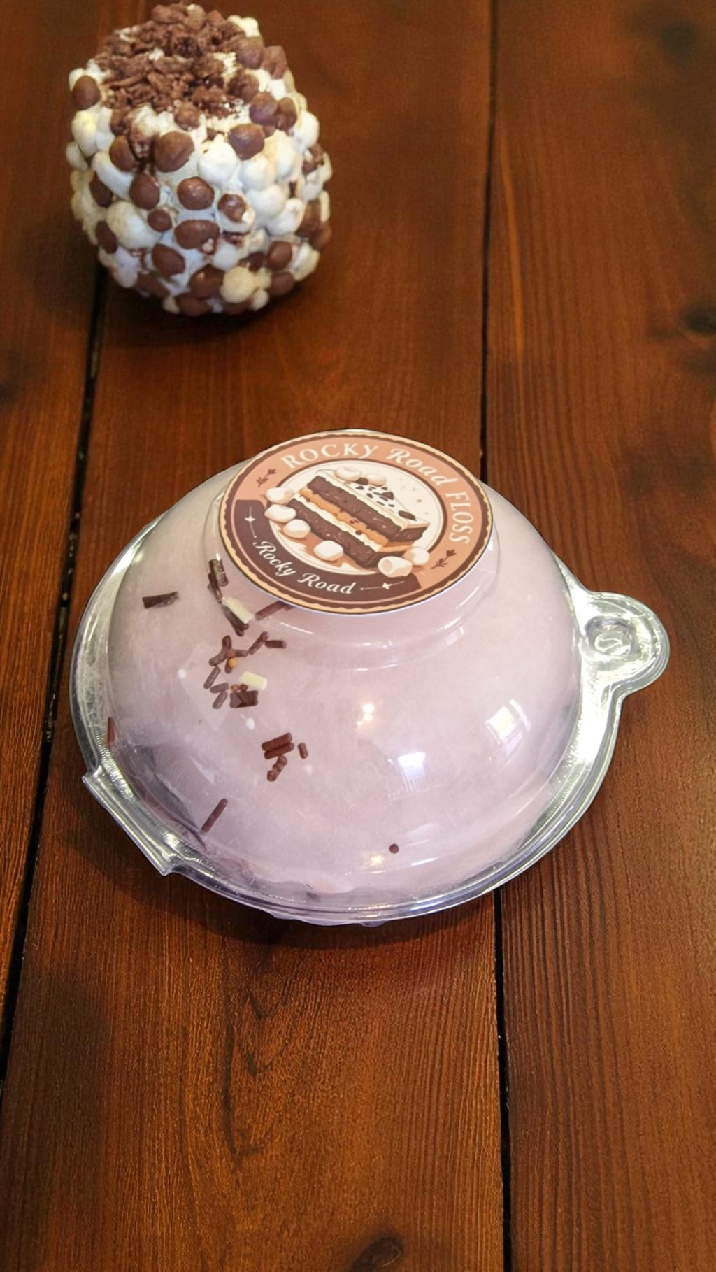 Pink dessert in a clear dome container on a wooden surface with a chocolate-covered treat in the background.