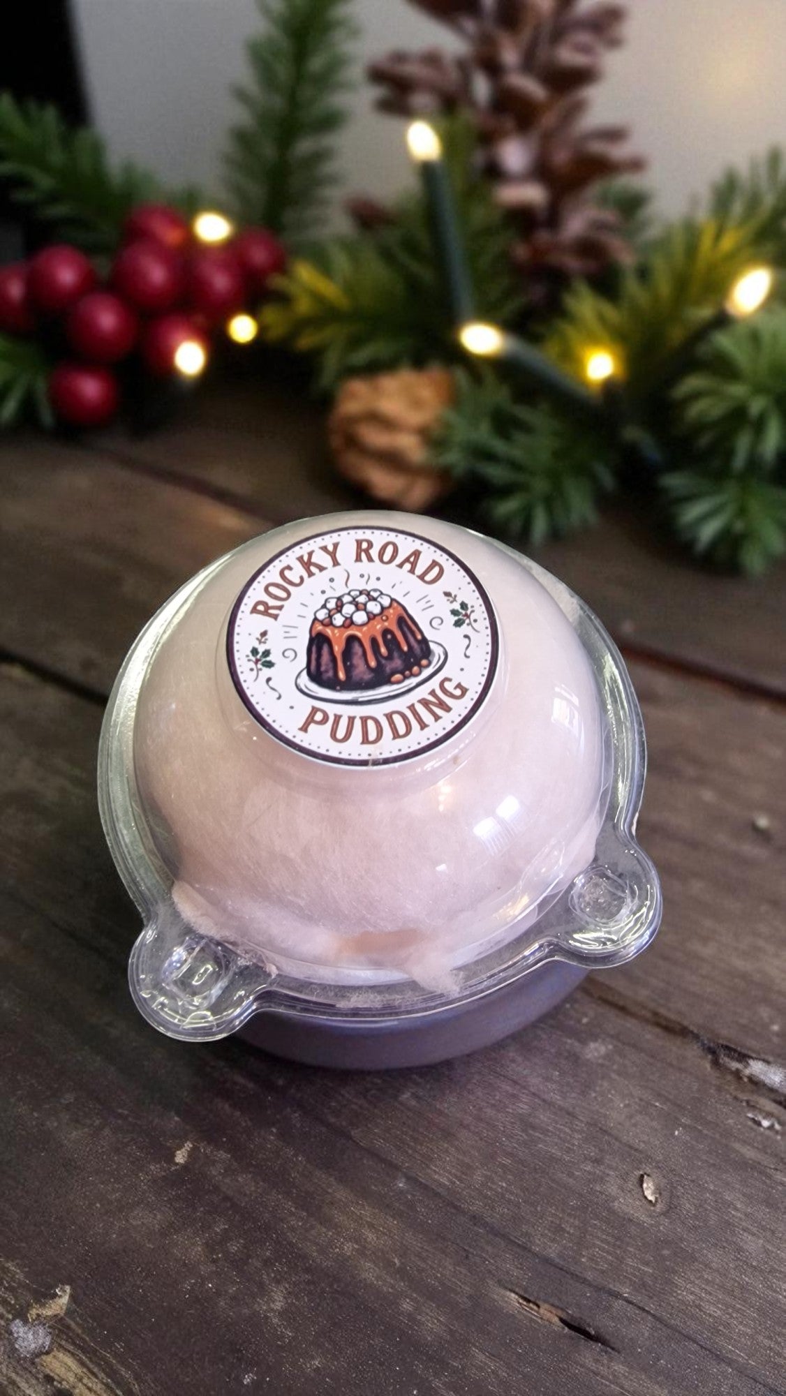 Bath bomb labeled 'Rocky Road Pudding' on a wooden surface with Christmas decorations in the background.