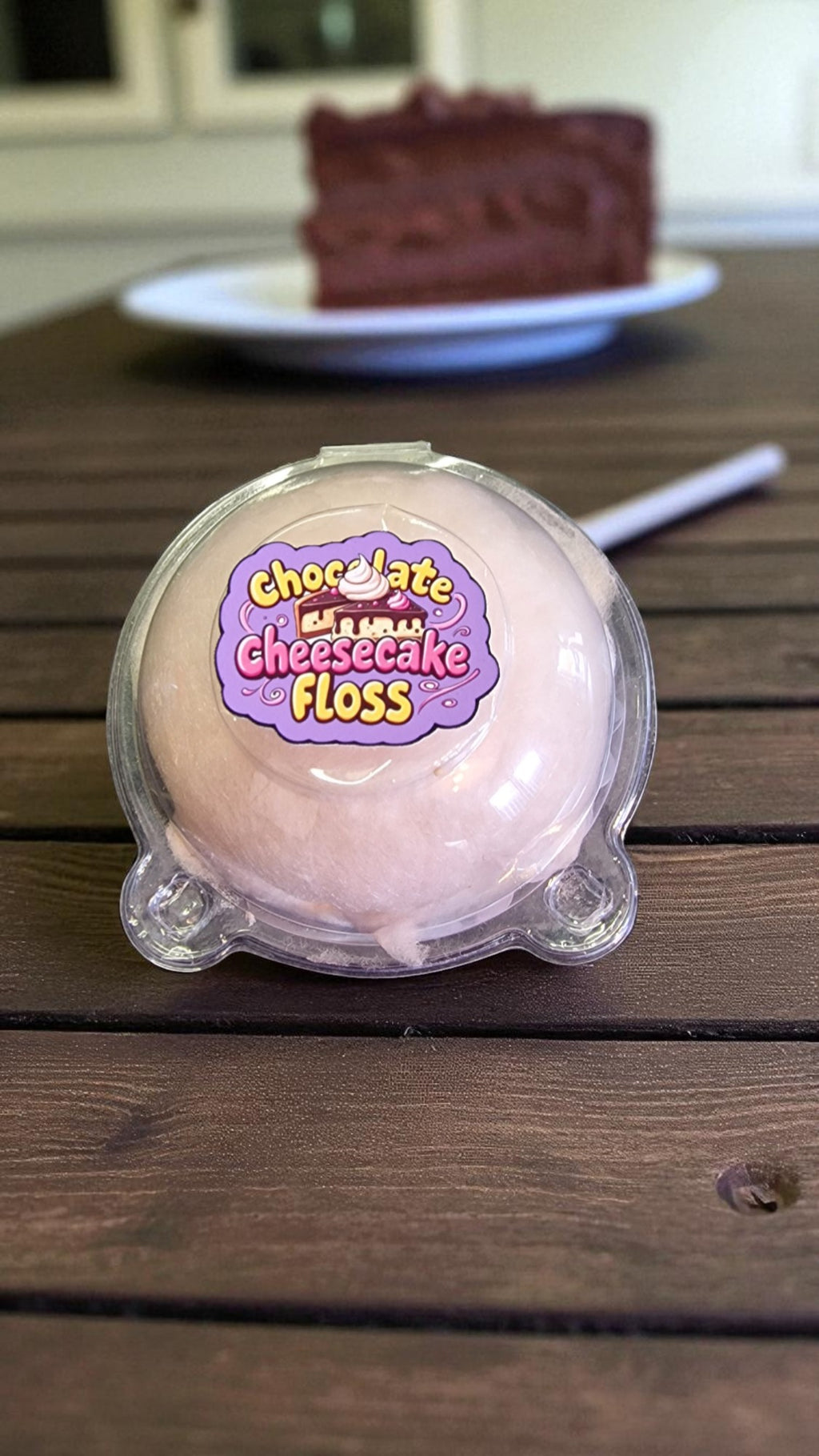 Pink floss ball with 'Chocolate Cheesecake Floss' label on a wooden table with a slice of chocolate cake in the background.