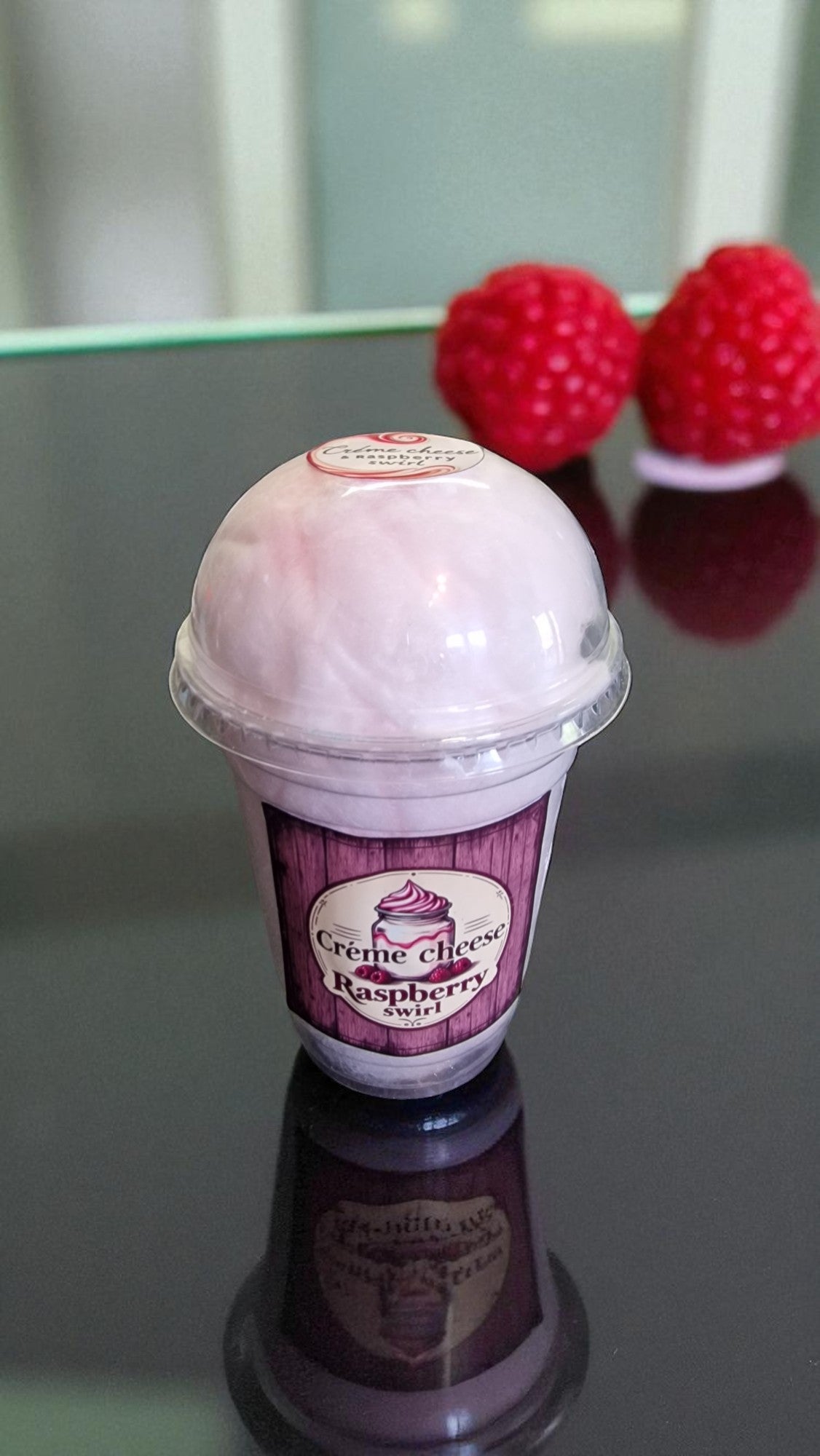Ice cream cone with raspberry swirl flavor label surrounded by raspberries on a reflective surface.