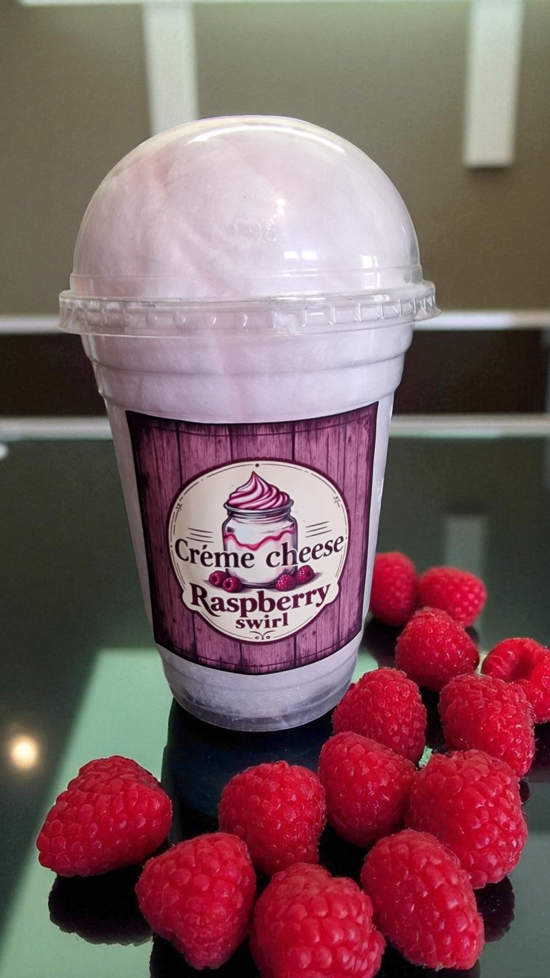 Ice cream cone with raspberry swirl flavor label surrounded by raspberries on a reflective surface.