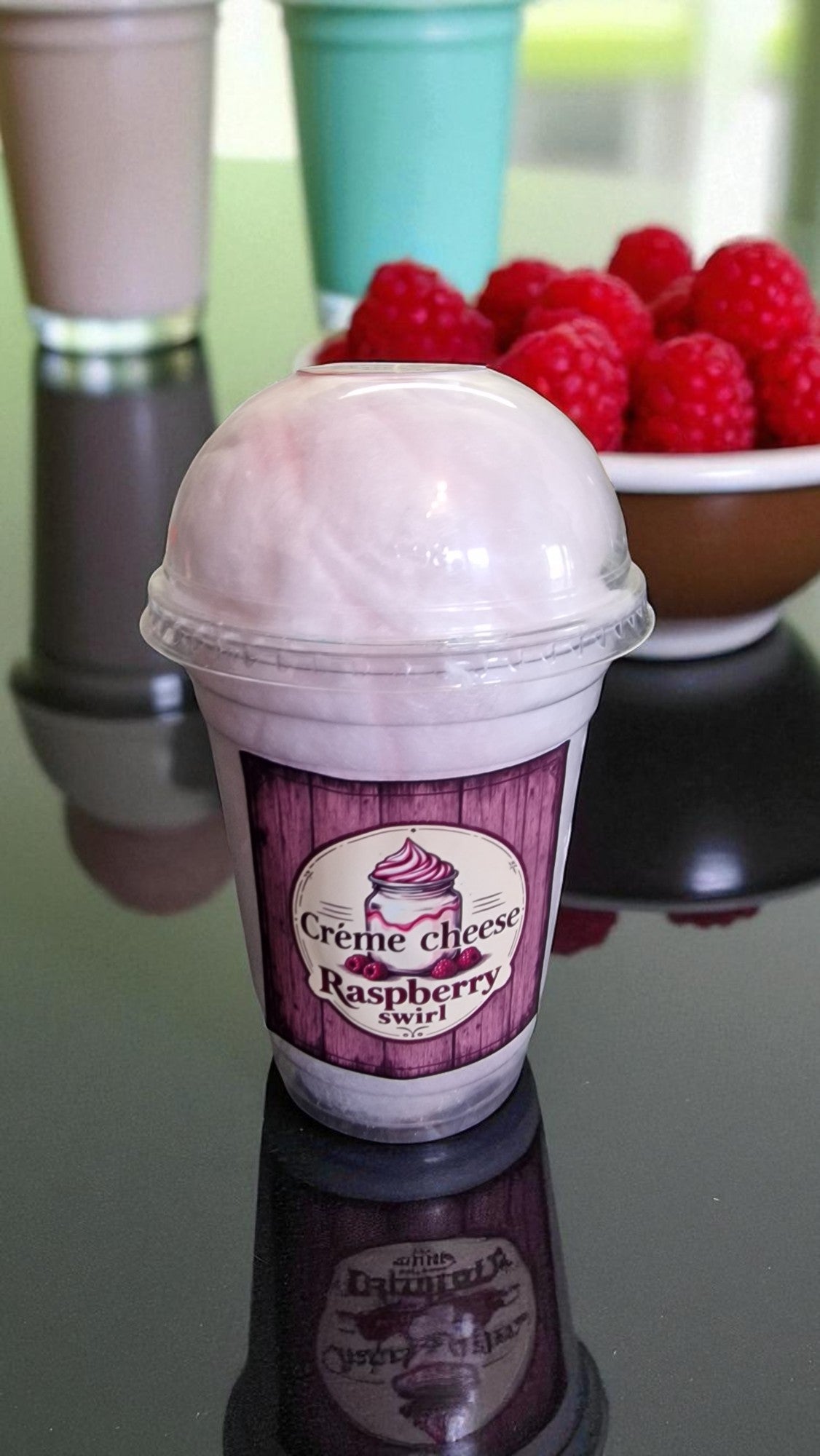 Ice cream shake with raspberry swirl flavor on a reflective surface, surrounded by raspberries and other drinks.