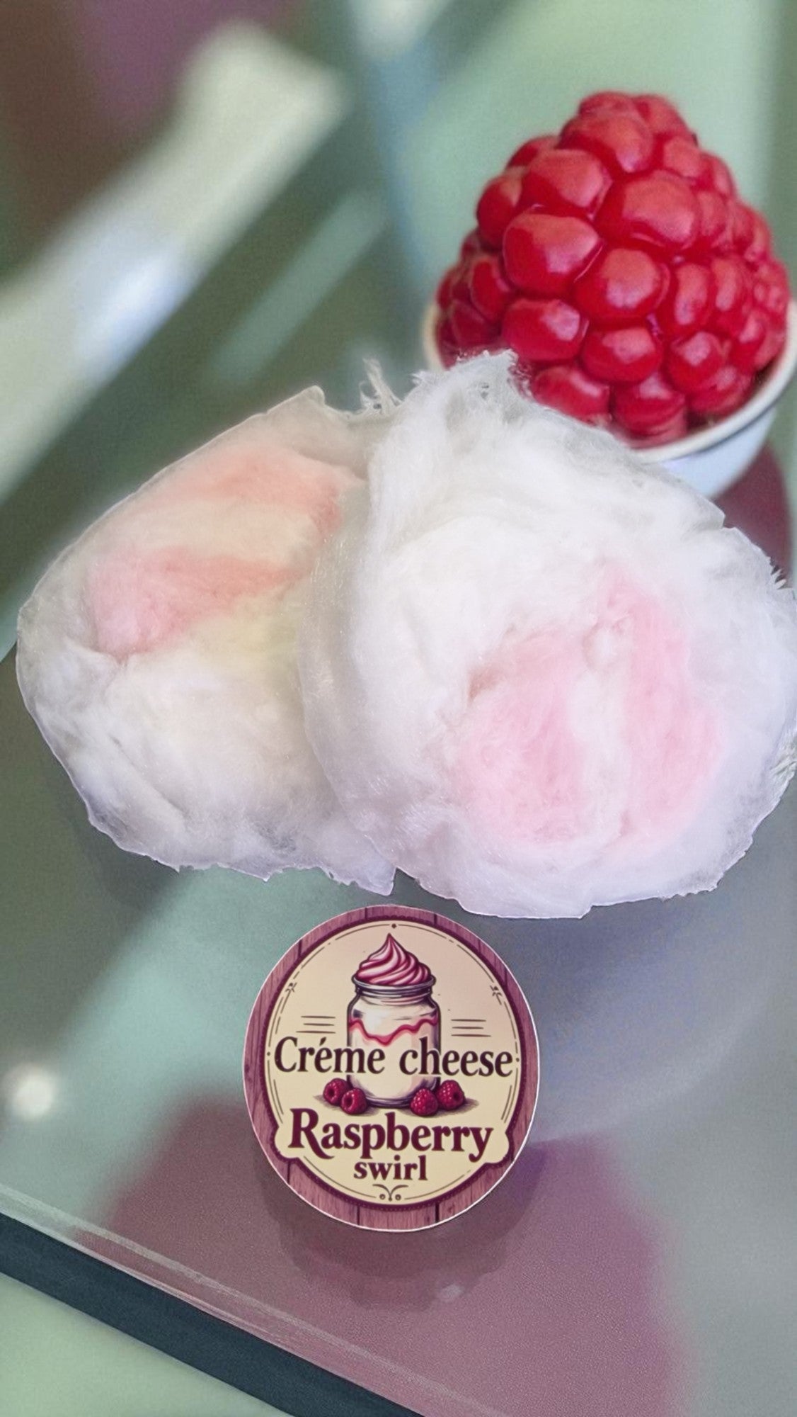 Two pieces of dessert with a raspberry on a reflective surface, featuring a 'Cèreme Cheese Raspberry Swirl' label.
