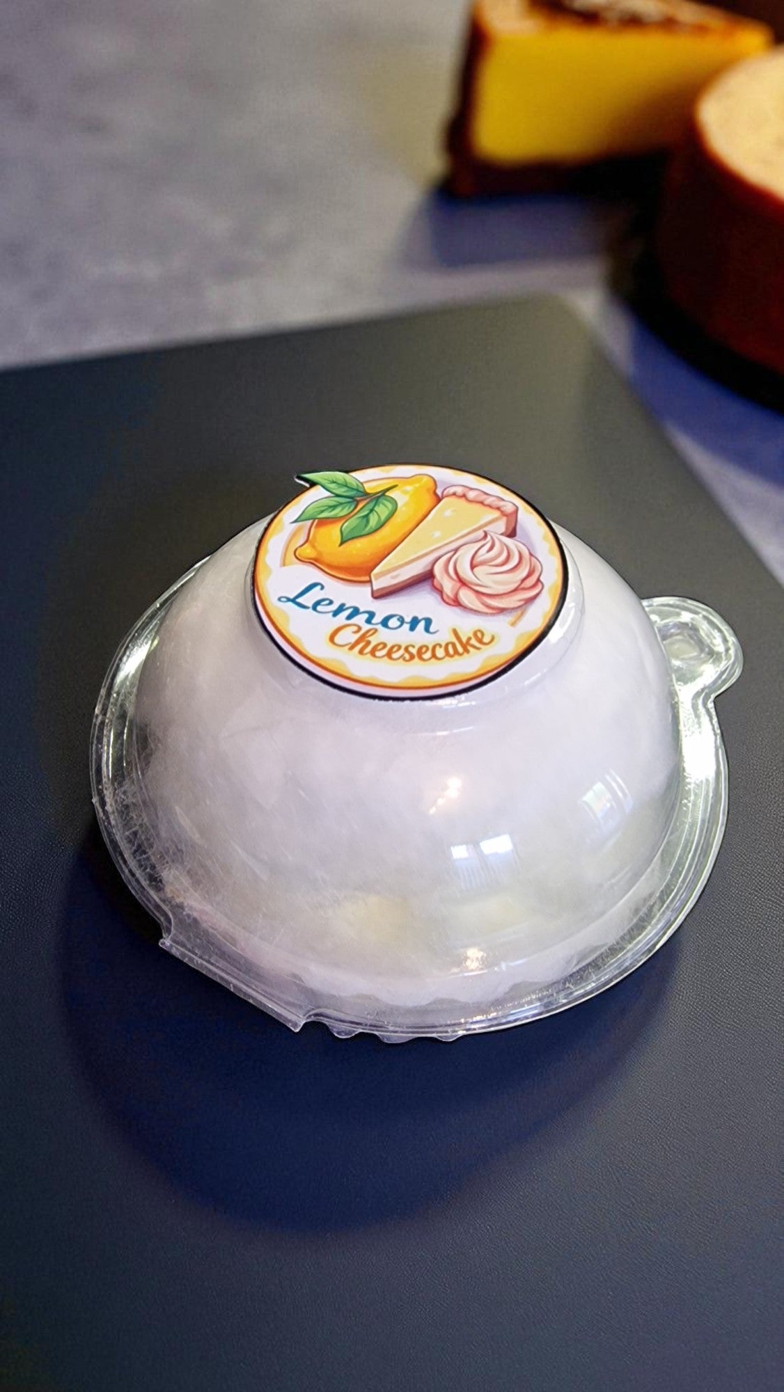 Lemon cheesecake in a clear plastic dome packaging on a gray surface.