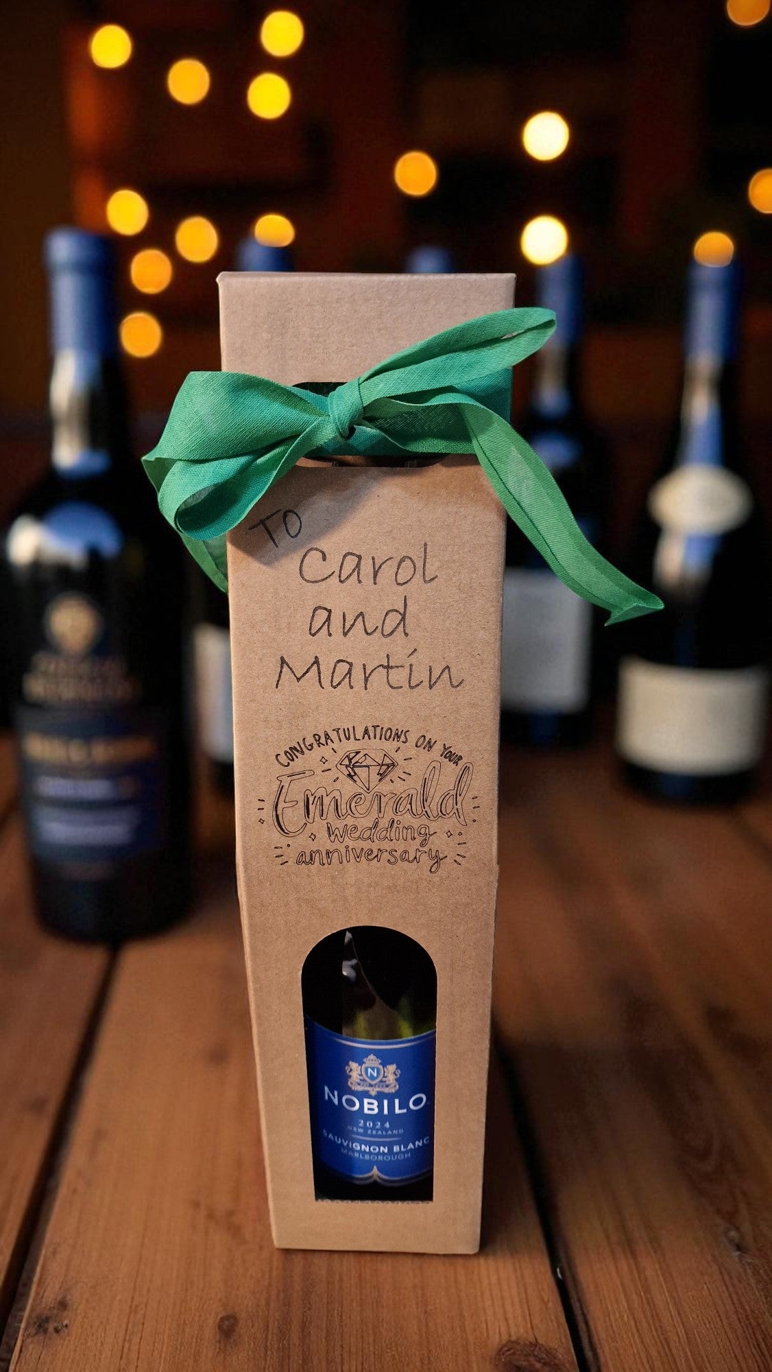 Wine bottle gift box with a green ribbon on a wooden surface with blurred wine bottles in the background.