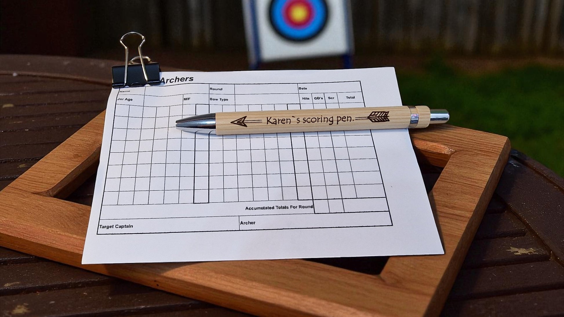 Scorecard with a pen on a wooden surface, with an archery target in the background.