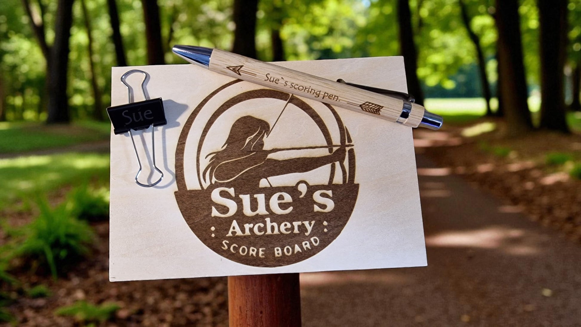 Sue's Archery Score Board with archery equipment on a wooden post outdoors.