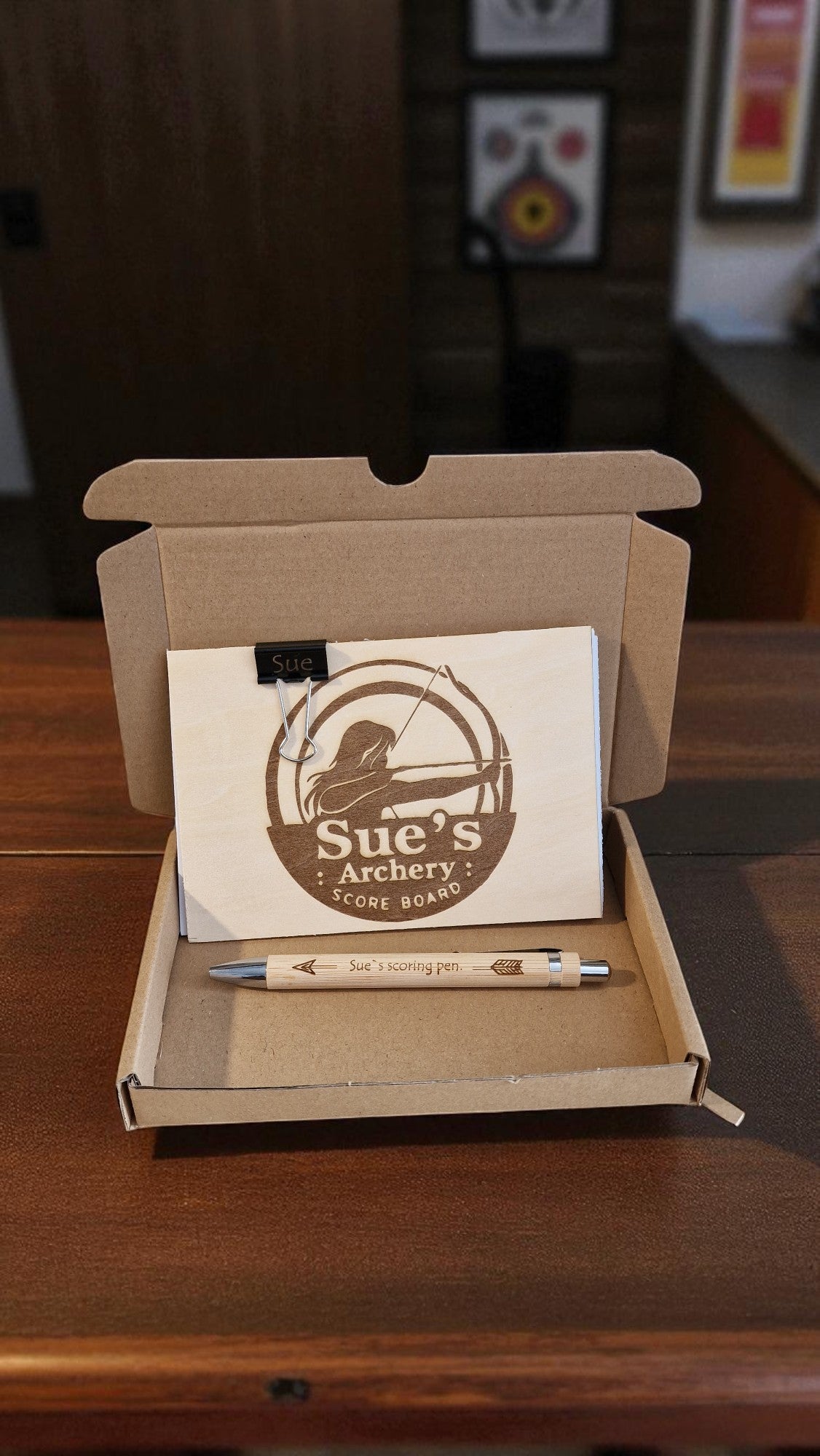 Box containing a notepad and pen with 'Sue's Archery' branding on a wooden surface.