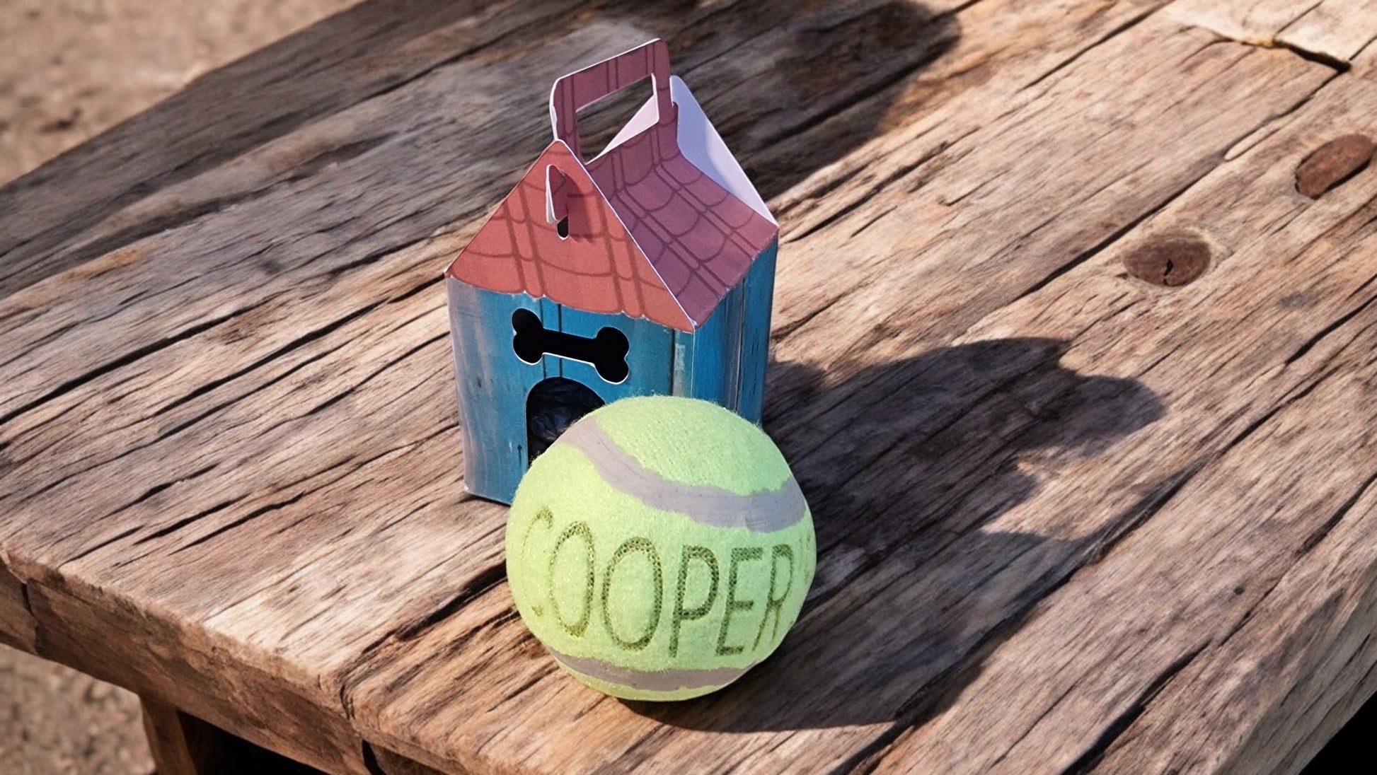 Tennis ball with 'COOPER' on a wooden surface next to a small dog house and bone-shaped toy.