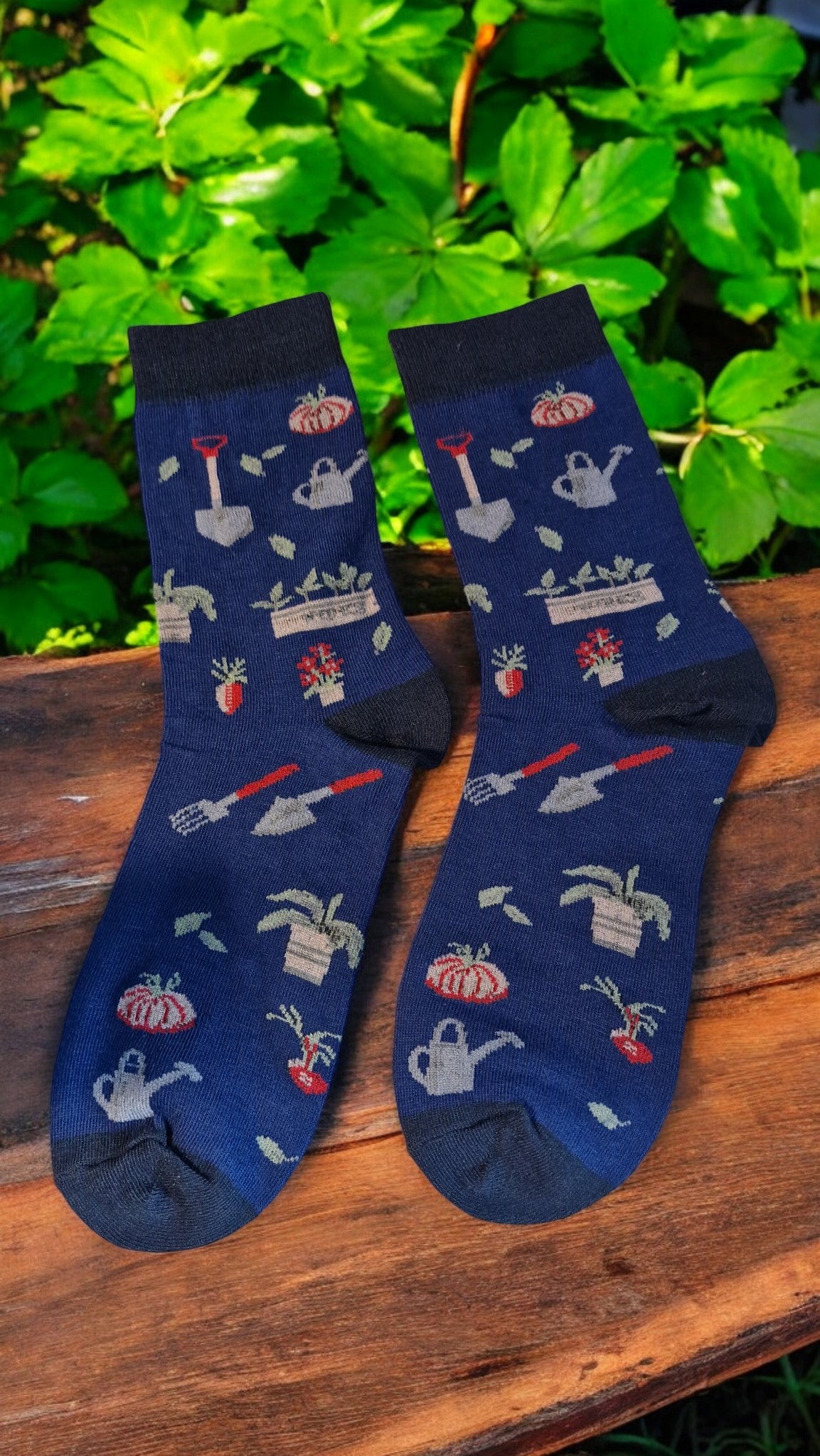 Pair of blue socks with gardening-themed designs on a wooden surface with green foliage in the background.