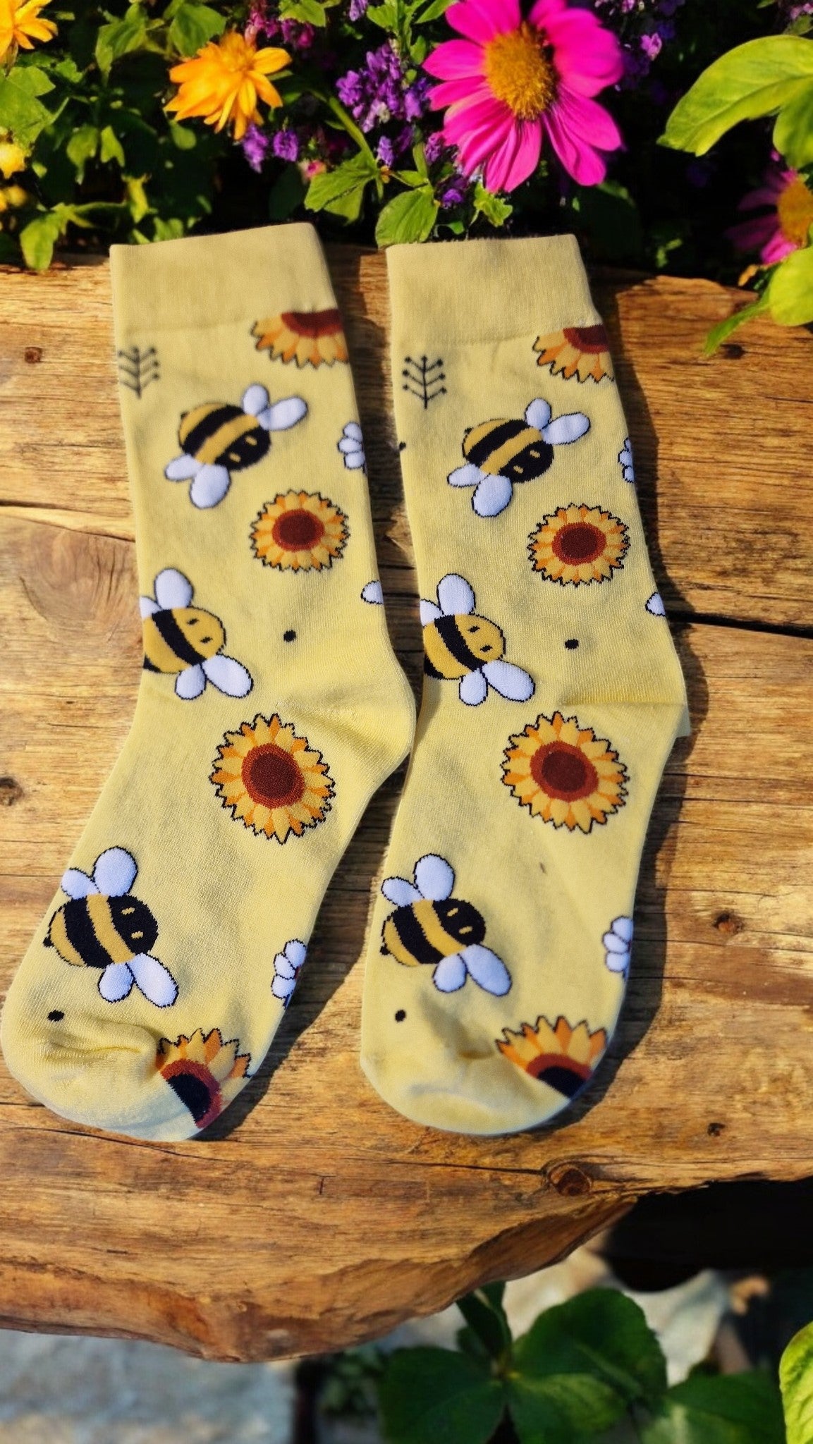 Beige socks with bee and sunflower pattern on a wooden surface with flowers in the background