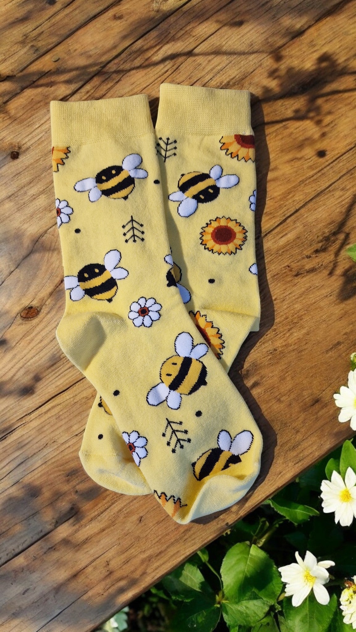 Yellow socks with bee and flower patterns on a wooden surface with flowers.