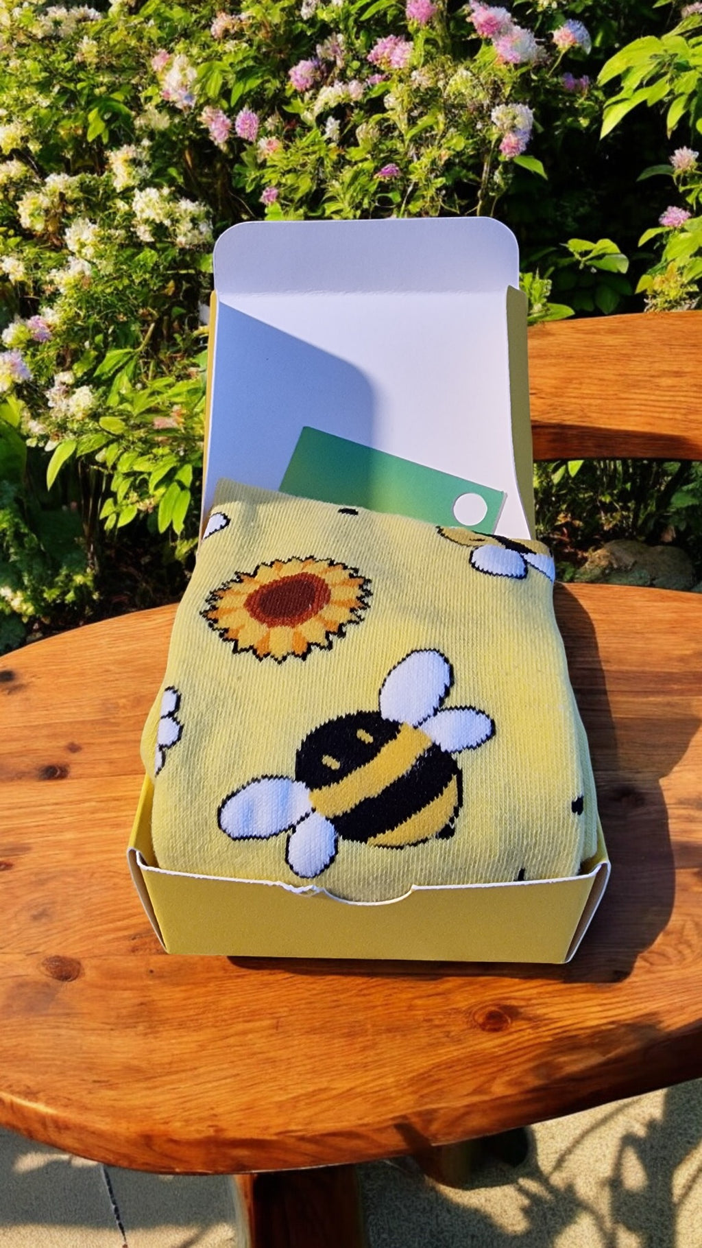 Socks with bee and sunflower design in a box on a wooden table outdoors.