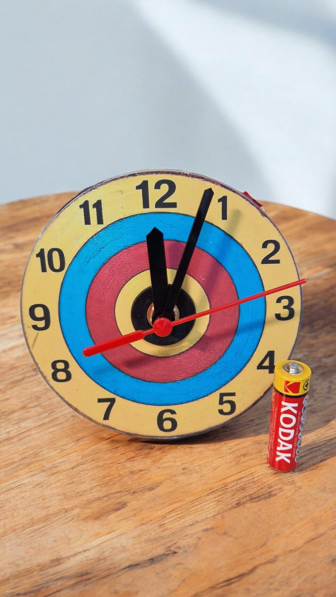 Colorful clock with a target design on a wooden surface, next to a Kodak battery.