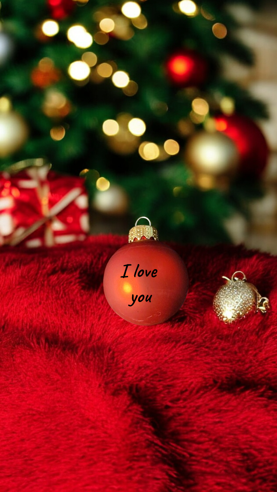 Red Christmas ornament with 'I love you' text on a red surface with a blurred Christmas tree in the background.