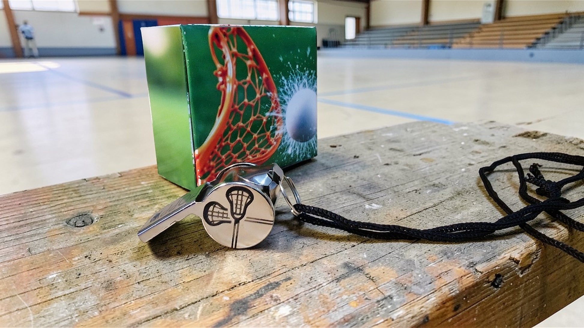 Lacrosse whistle with a packaging box on a wooden surface in an indoor sports facility.