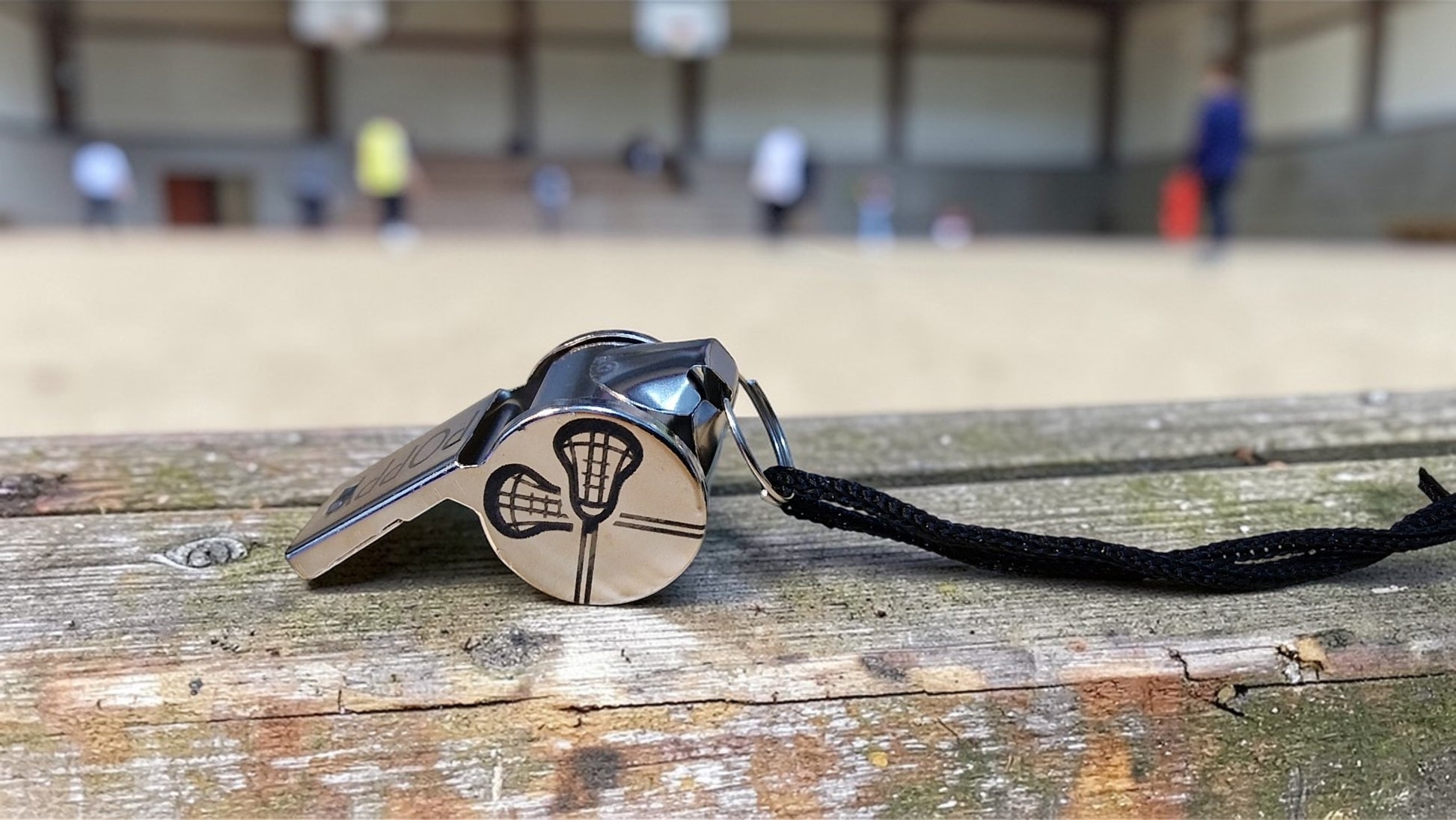 Metallic whistle with a black lanyard on a wooden surface, with a blurred sports field background.
