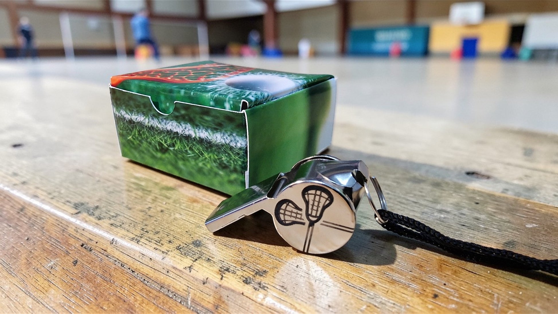 Green lacrosse box and silver whistle with black lanyard on a wooden surface.