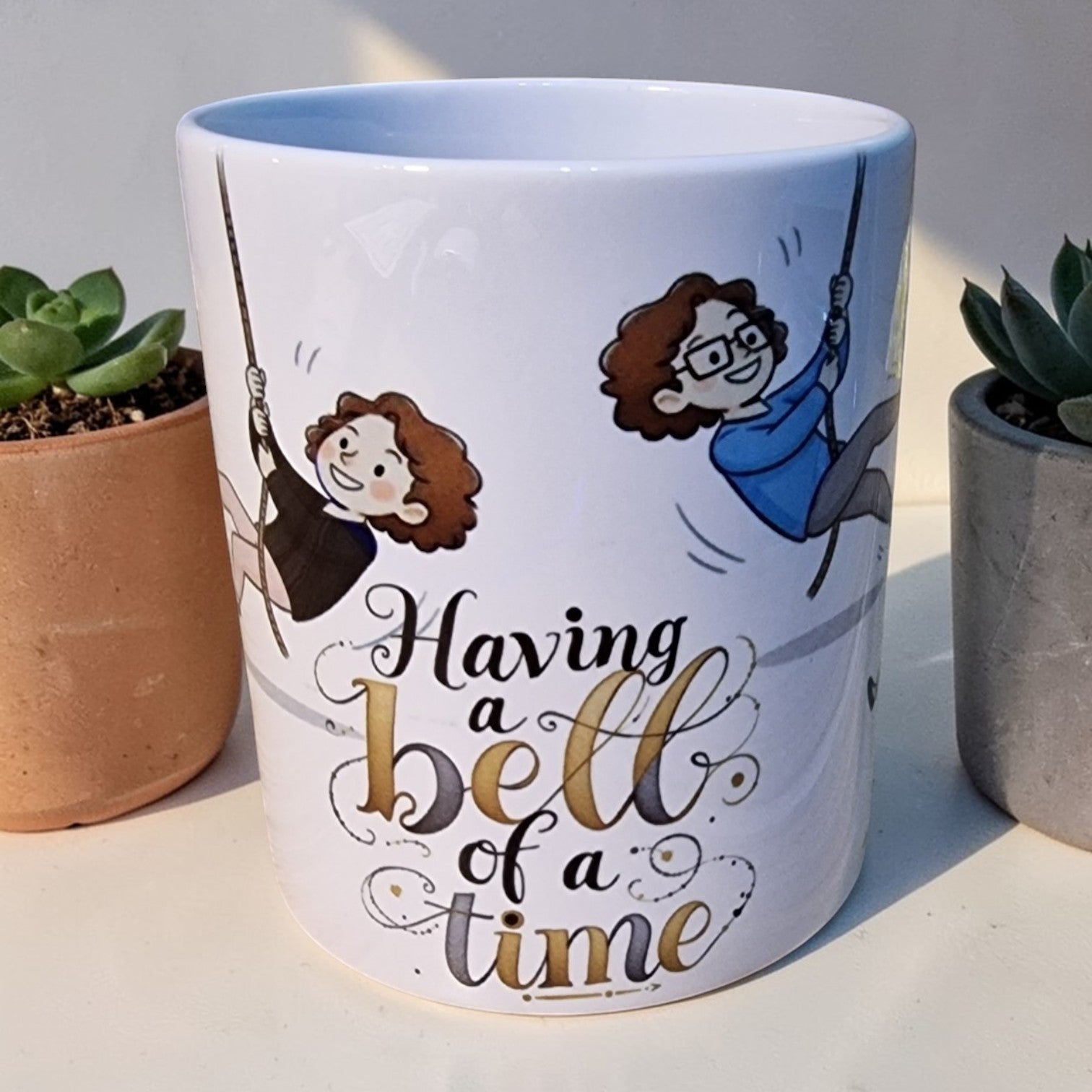 Mug with illustrations of two people on a swing and text 'Having a bell of a time' on a light background.
