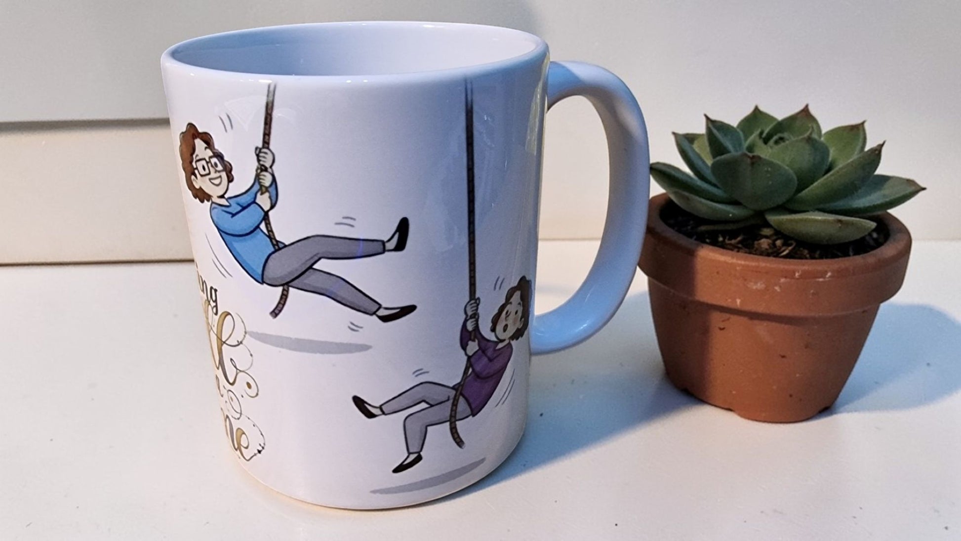 Mug with a design of two people on swings next to a potted plant on a white surface
