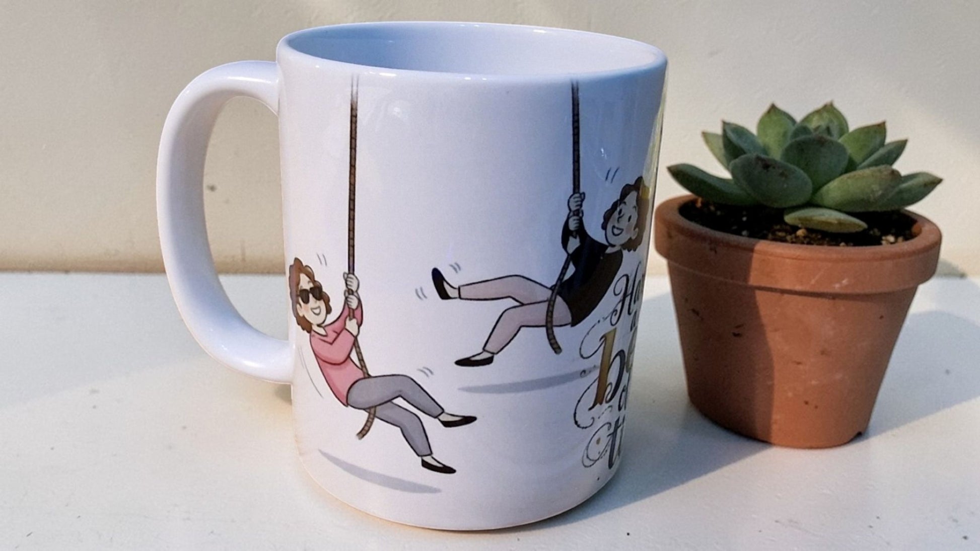 White mug with illustrations of a man and woman on swings, next to a potted plant.