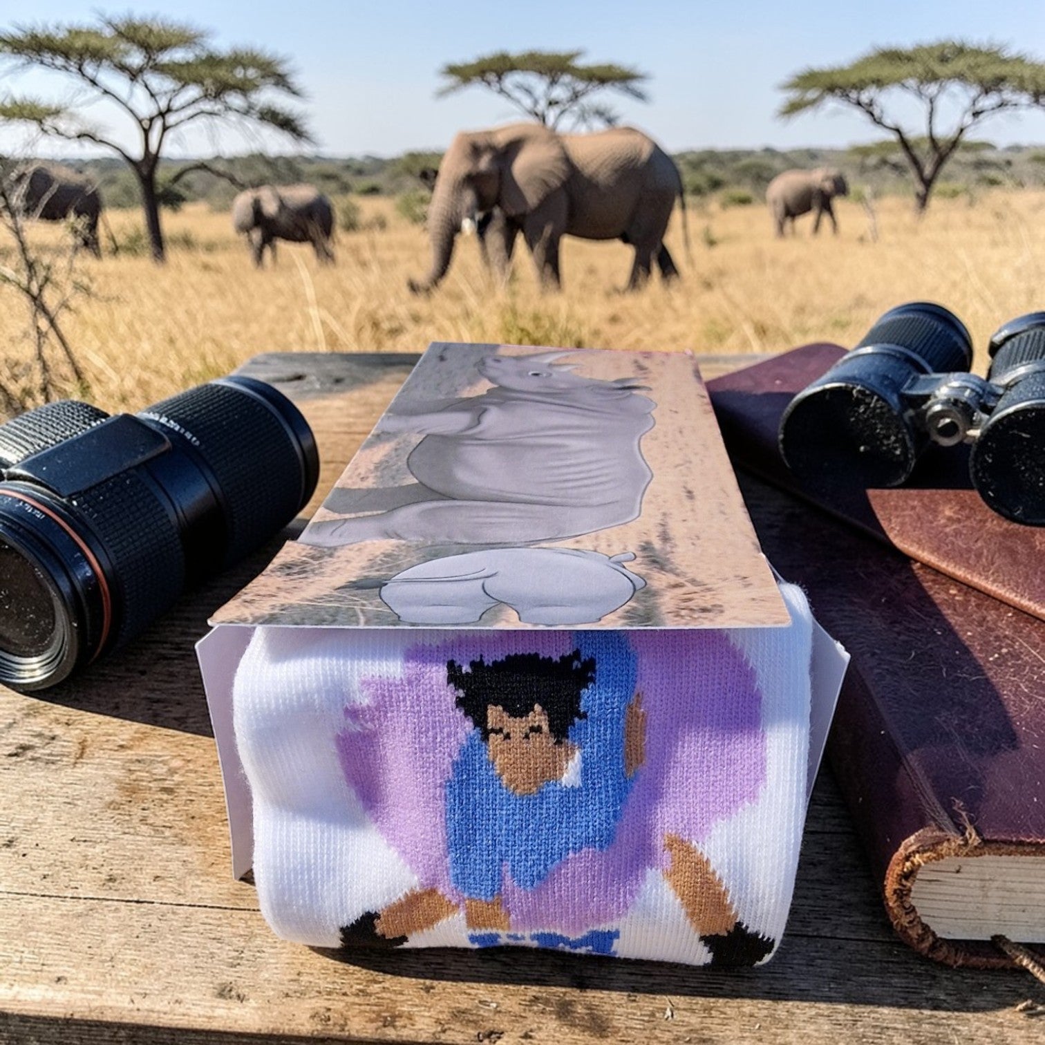 Socks with a design of a person with braids and a purple shirt on a wooden surface with binoculars and a book, with elephants in the background.