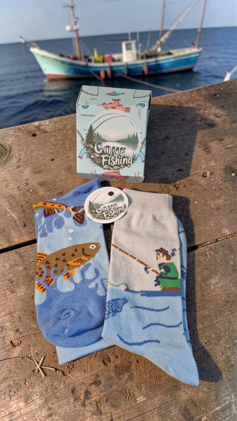 Fishing-themed socks with a box labeled 'Game Fishing' on a wooden surface with a boat in the background.