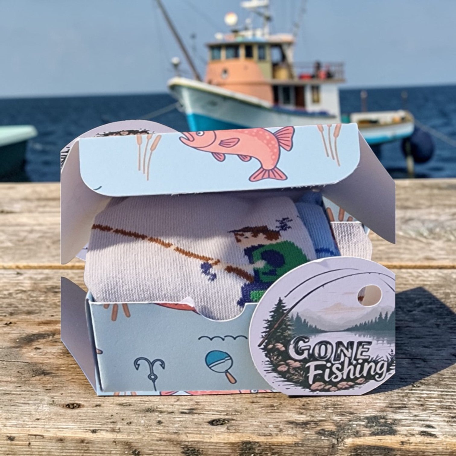 Box of socks with a 'Gone Fishing' design on a wooden surface with a boat in the background.