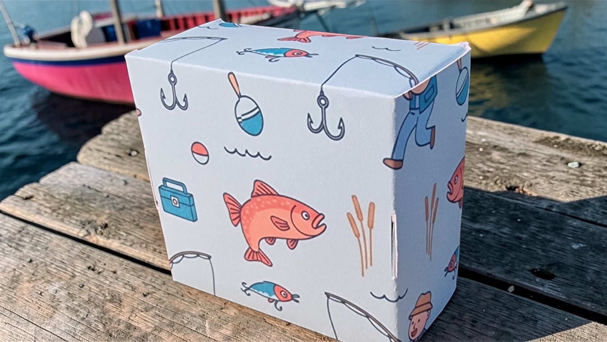 Box with fishing-themed illustrations on a wooden dock with boats in the background