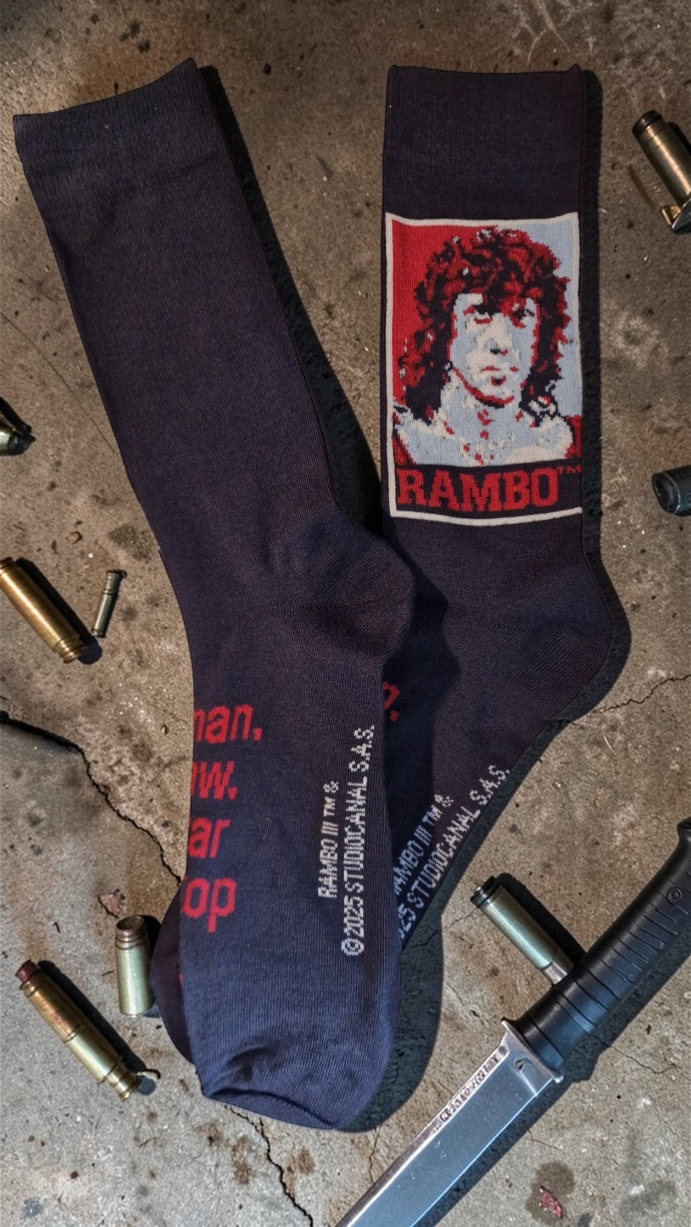 Black socks with a graphic design of a face and 'RAMBO' text on a concrete surface with gun parts.