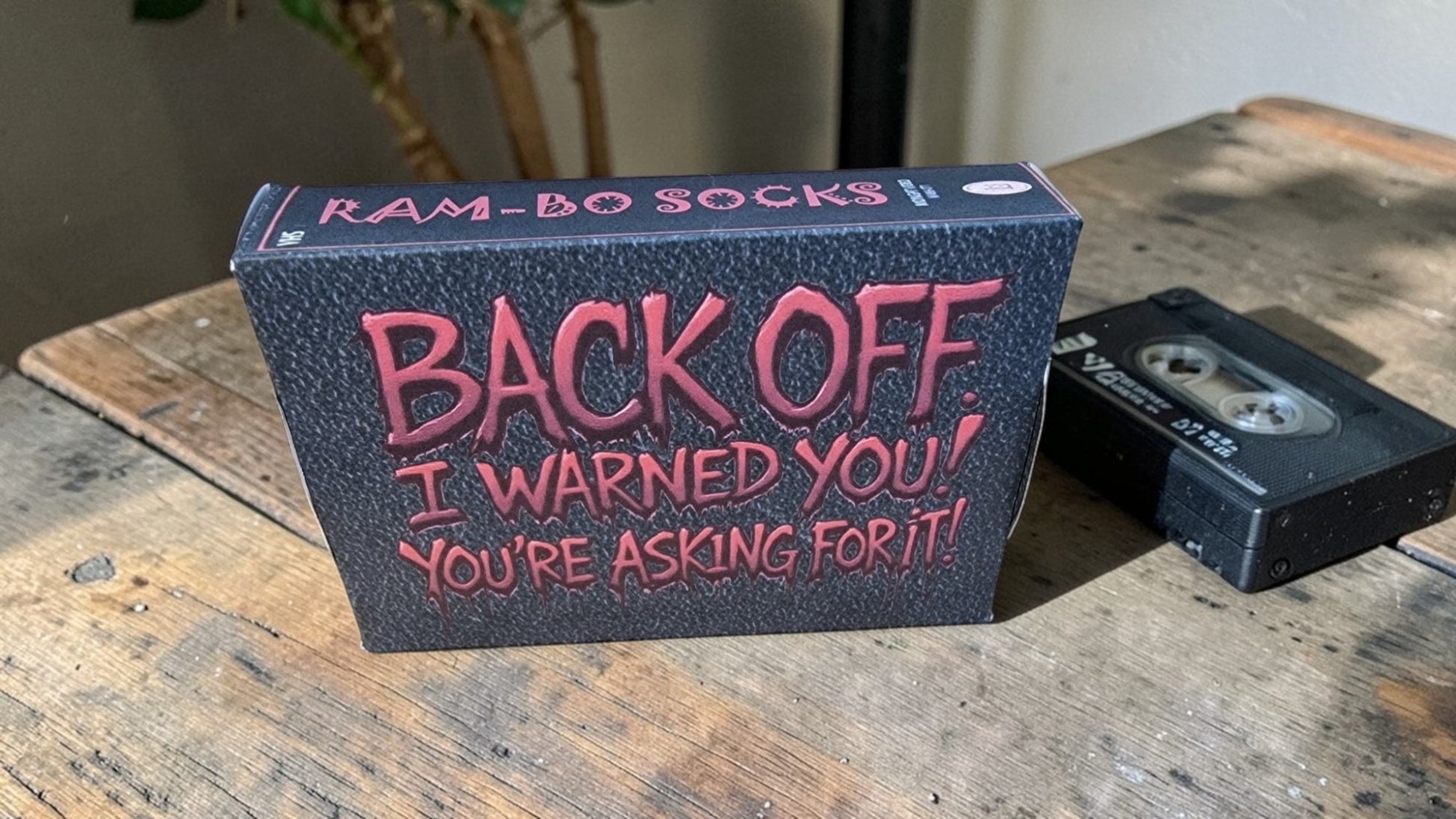 Box of 'Ram-Bo Socks' with humorous text on a wooden surface with a cassette tape.