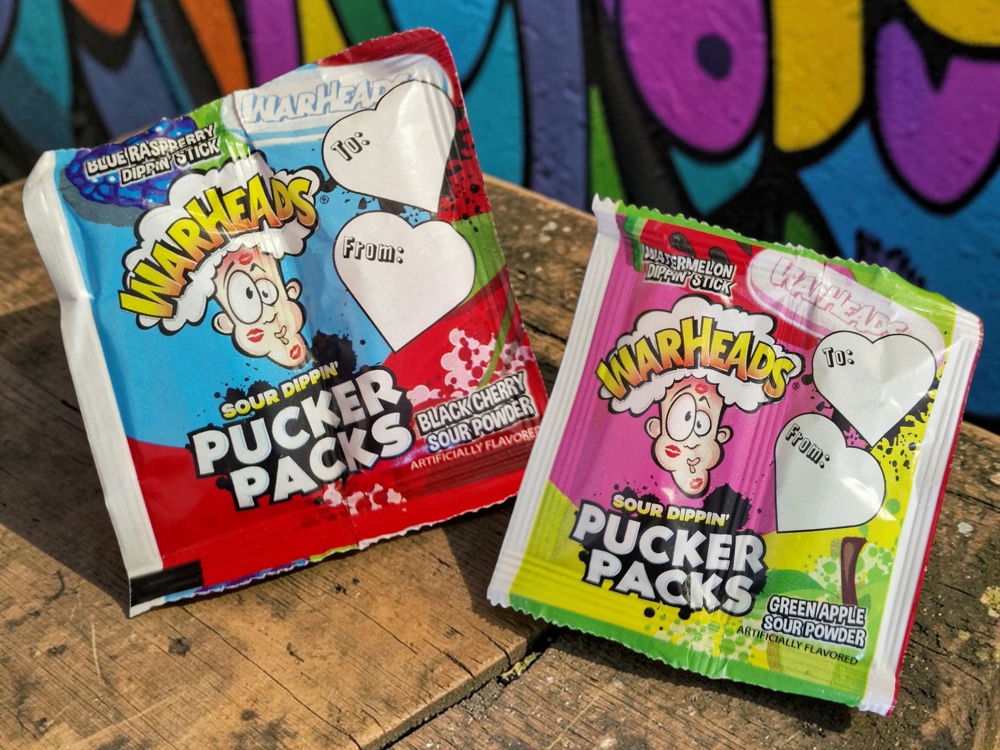 Two packages of Warheads Pucker Packs on a wooden surface with a colorful graffiti background.