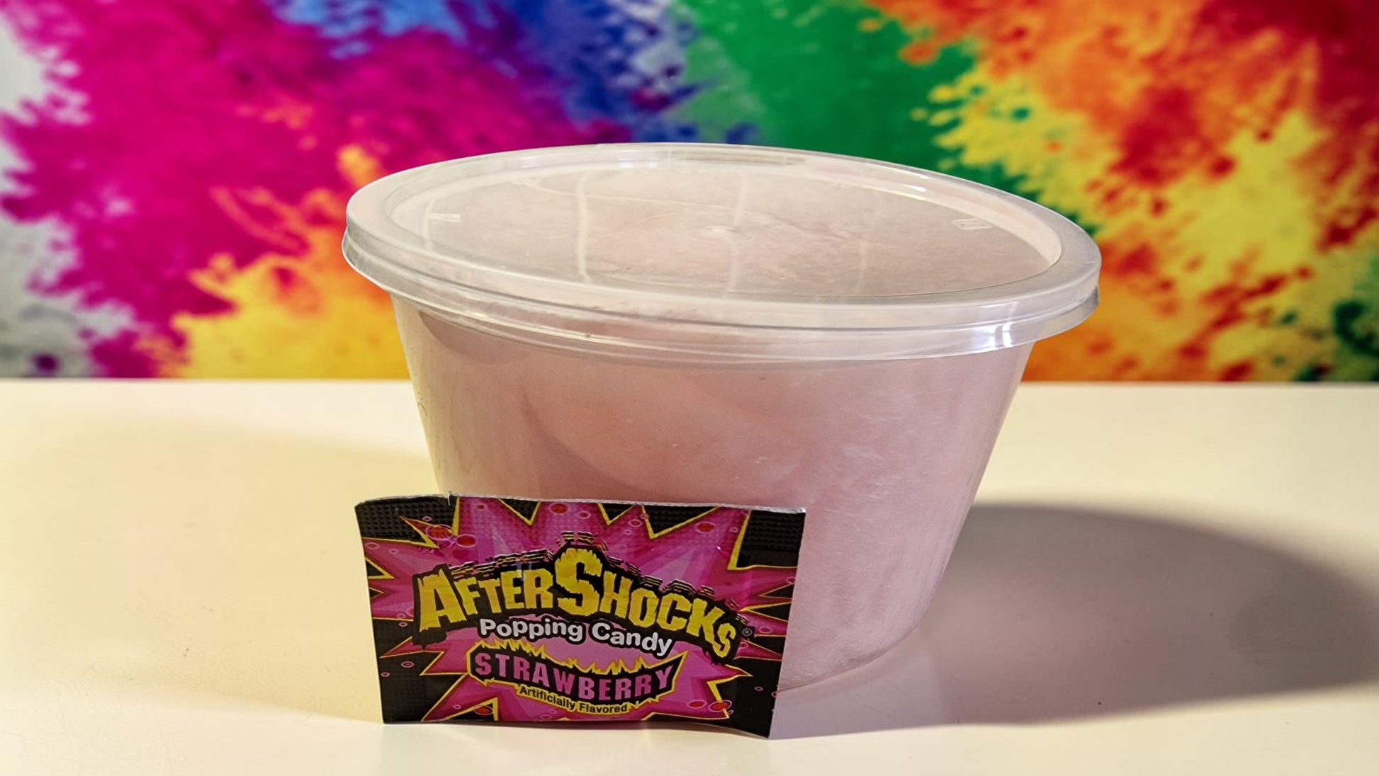Pink container of AfterShock Popping Candy Strawberry with colorful tie-dye background