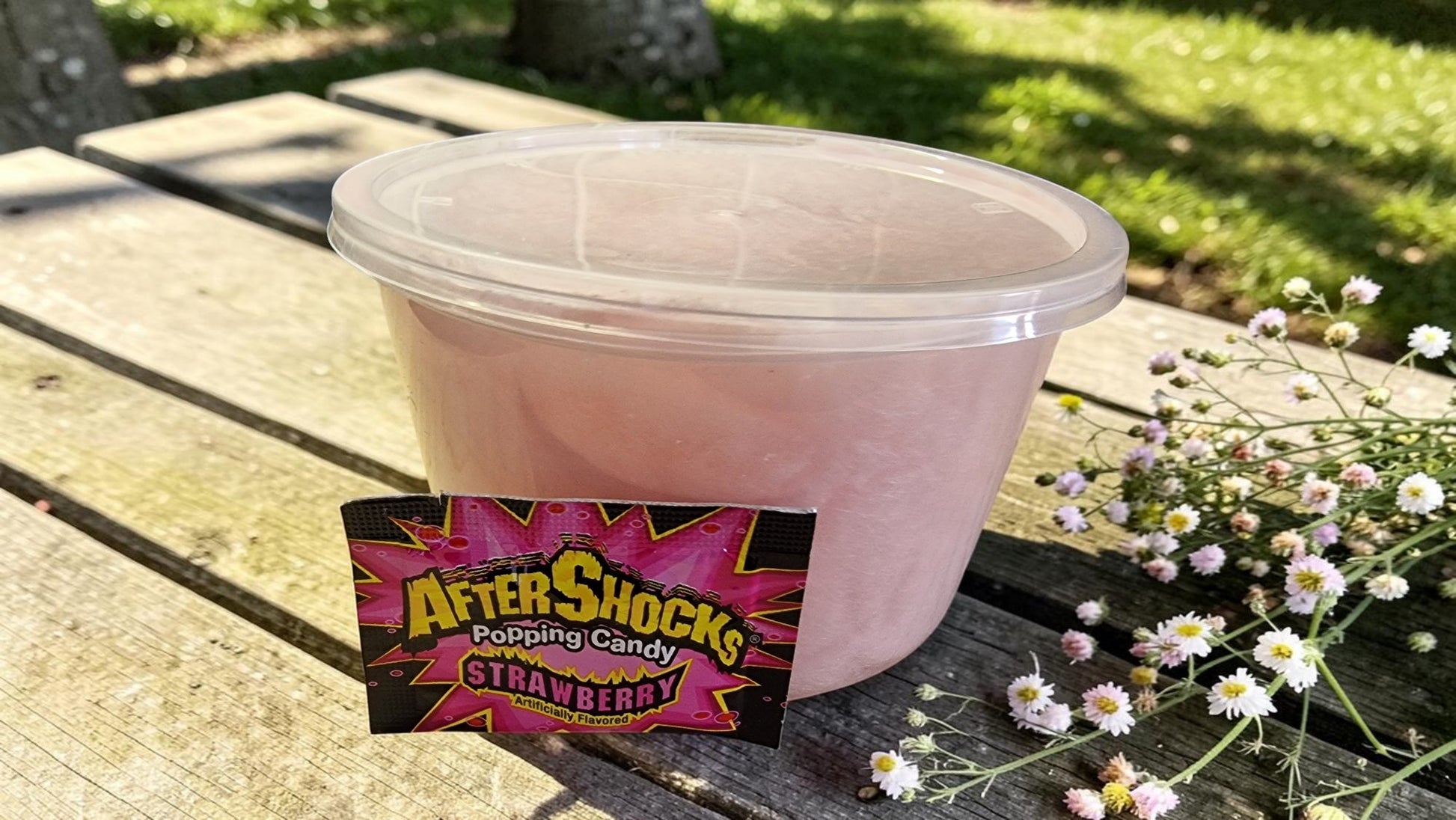 Pink container of AfterShocks popping candy on a wooden surface with flowers and grass in the background