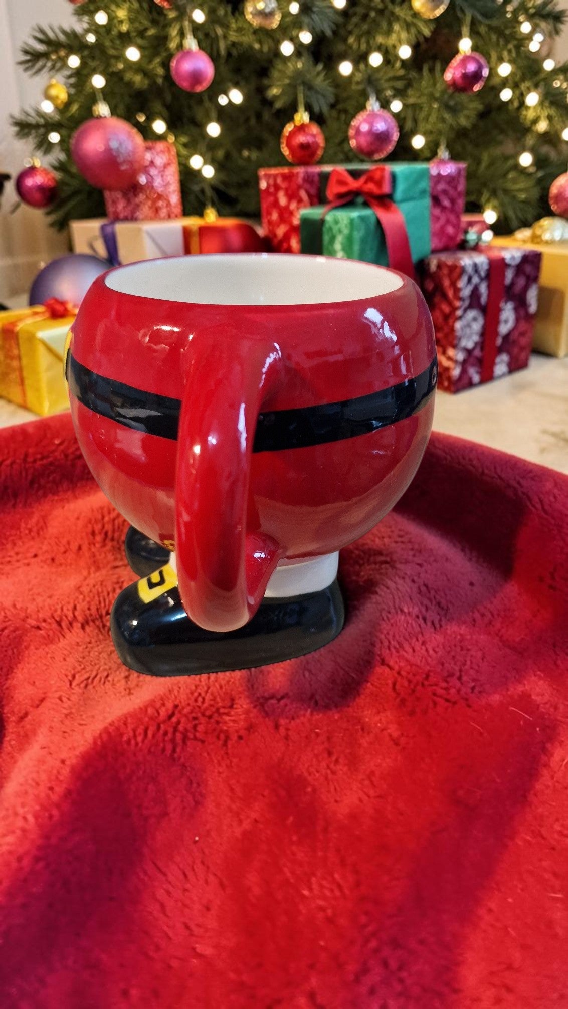 Red mug with black handle and base on a red surface, Christmas tree and presents in the background