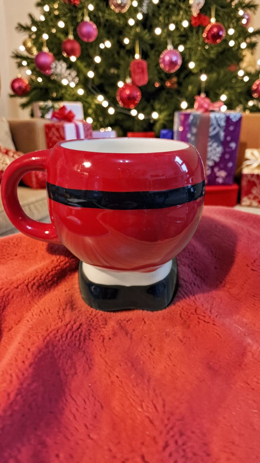 Red mug with black stripe on a red blanket in front of a decorated Christmas tree.