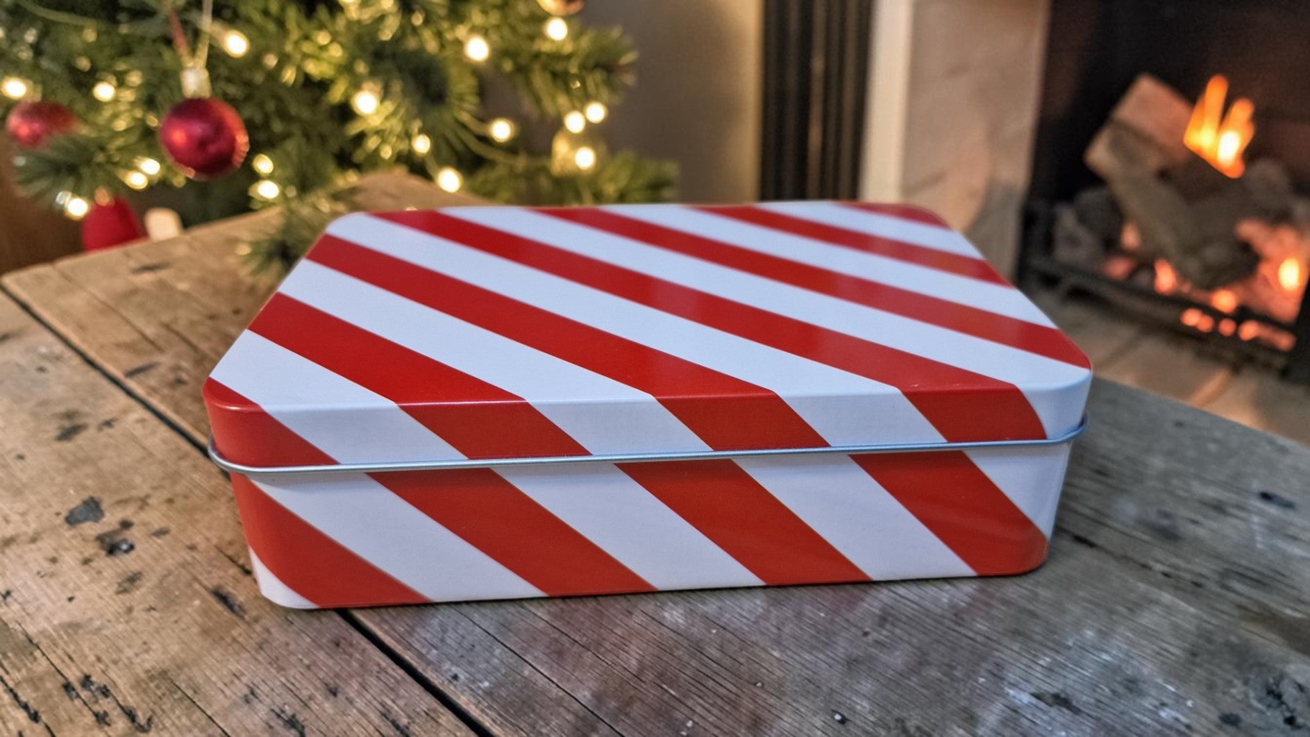Red and white striped box on a wooden table with a Christmas tree and fireplace in the background