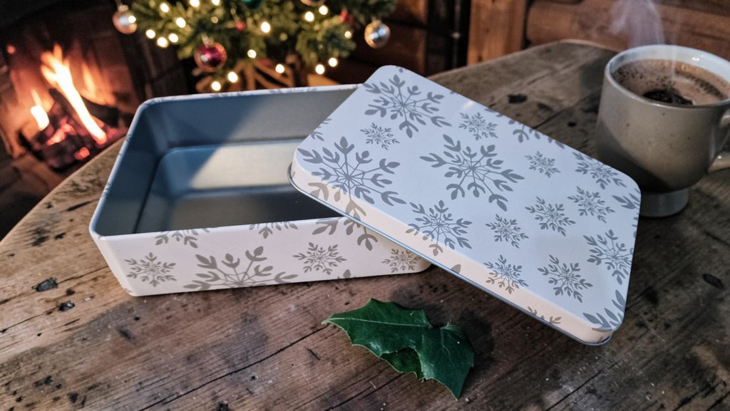 Decorative tin box with floral lid on a wooden table with a cup of coffee and Christmas tree in the background.