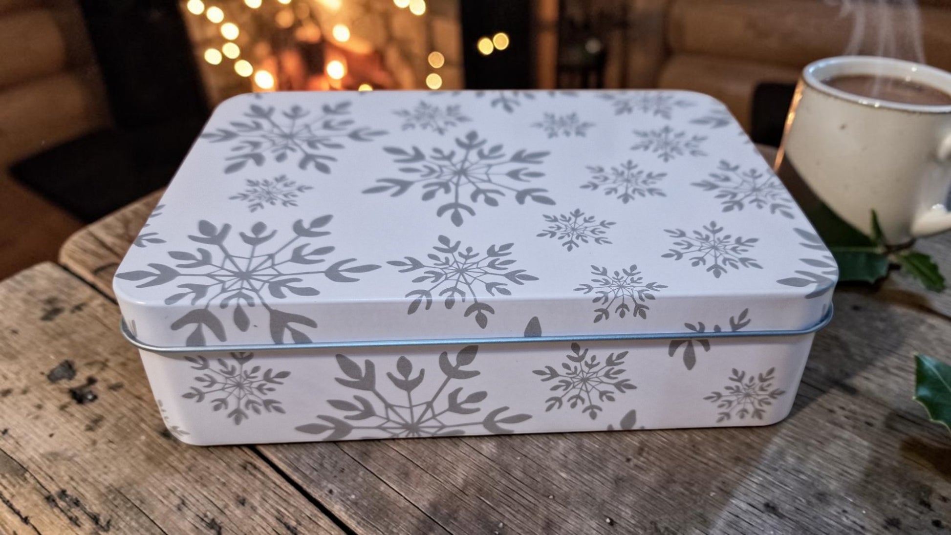 Decorative tin box with snowflake pattern on a wooden surface, with a blurred background of lights and a cup.