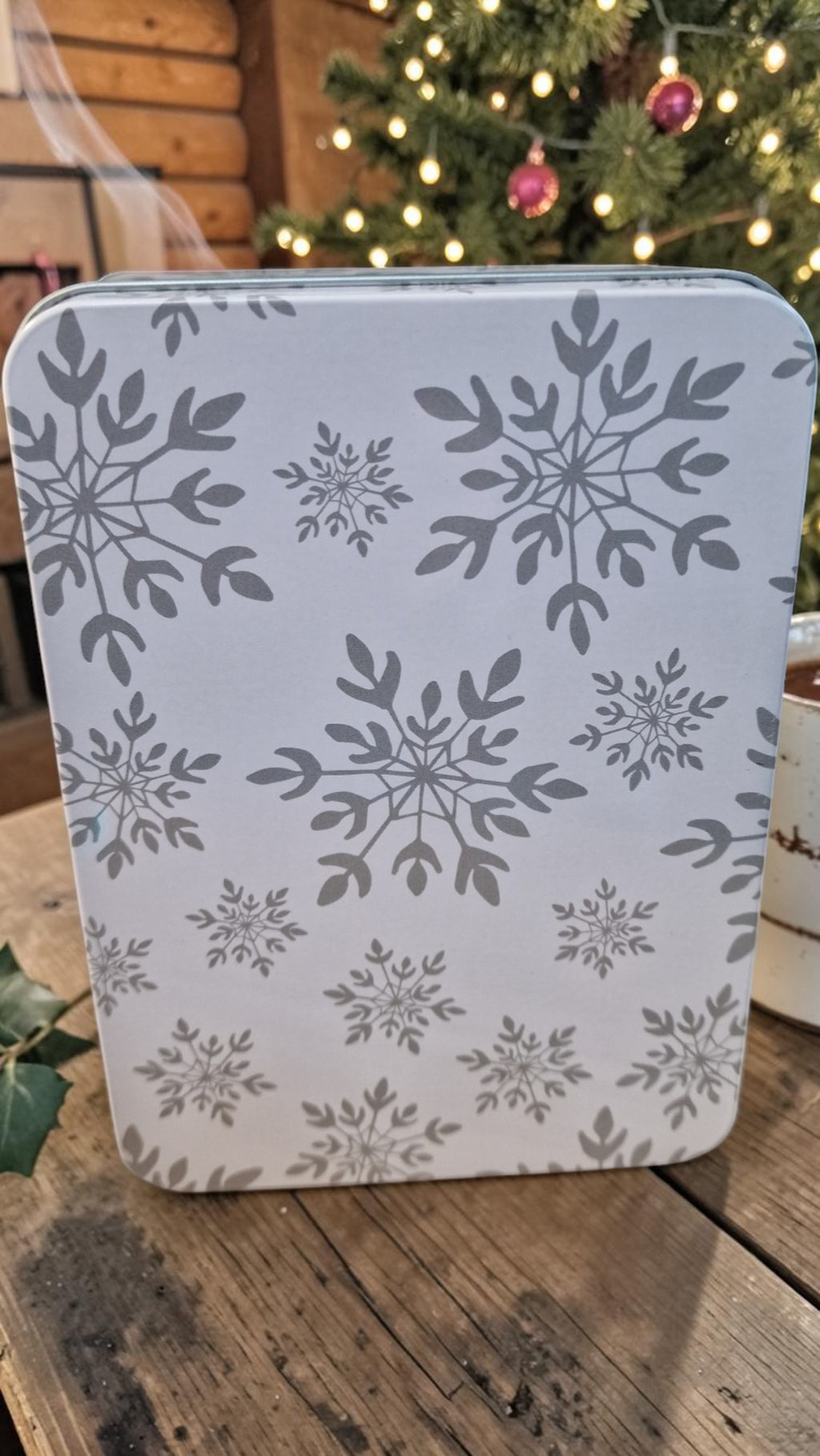 White container with gray snowflake pattern on a wooden surface with a Christmas tree in the background
