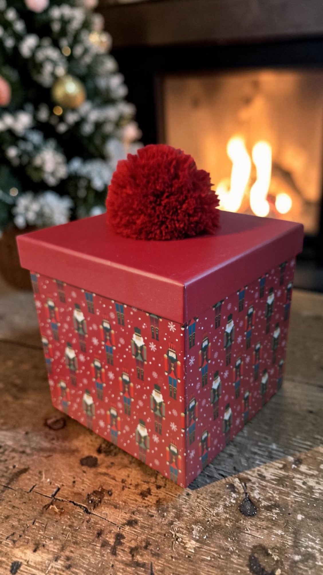 Red gift box with a decorative top on a wooden surface, Christmas tree and fireplace in the background