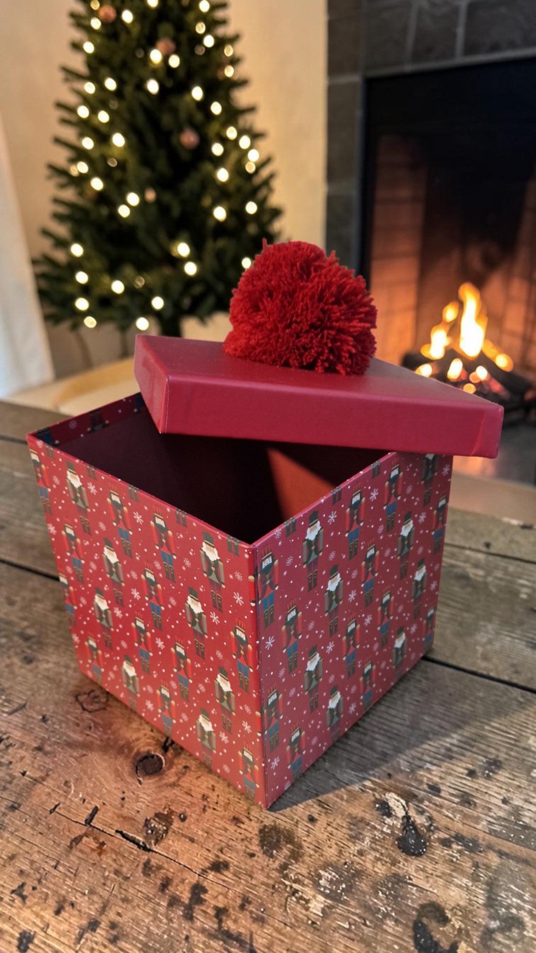Red gift box with a red pom-pom on top, placed on a wooden surface with a Christmas tree and fireplace in the background.