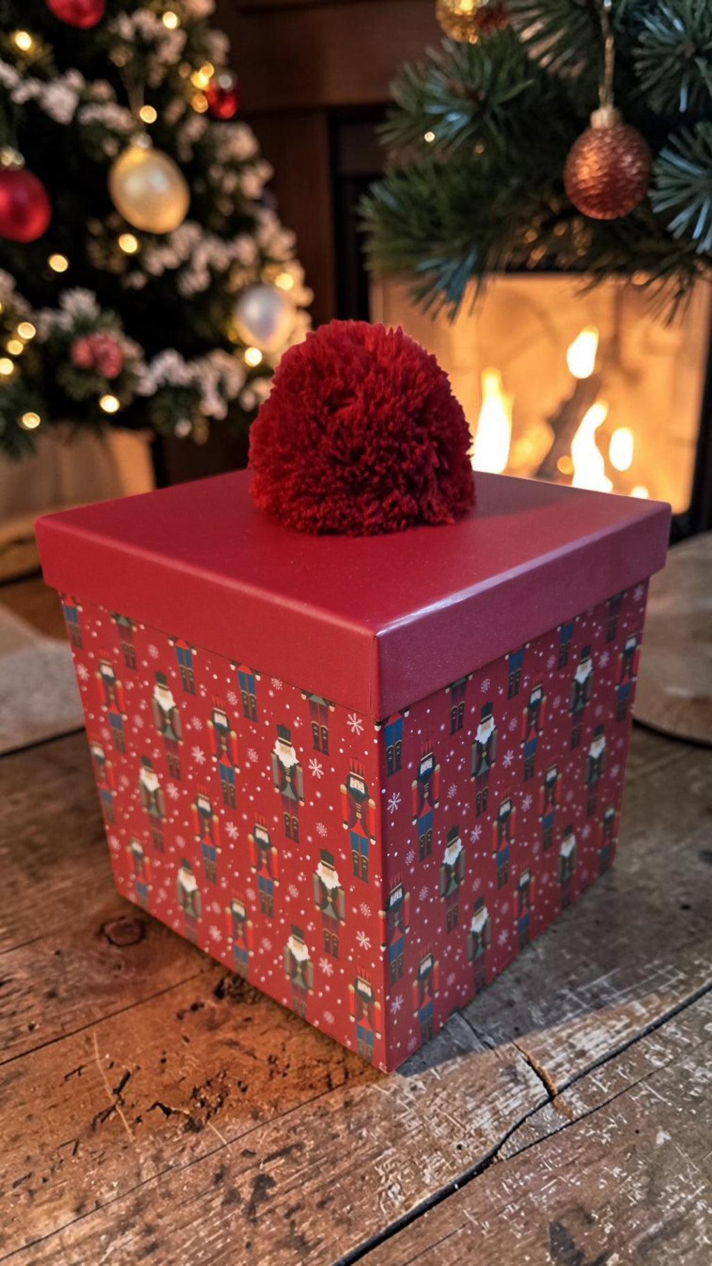Red gift box with a decorative red ball on top, Christmas tree and fireplace in the background