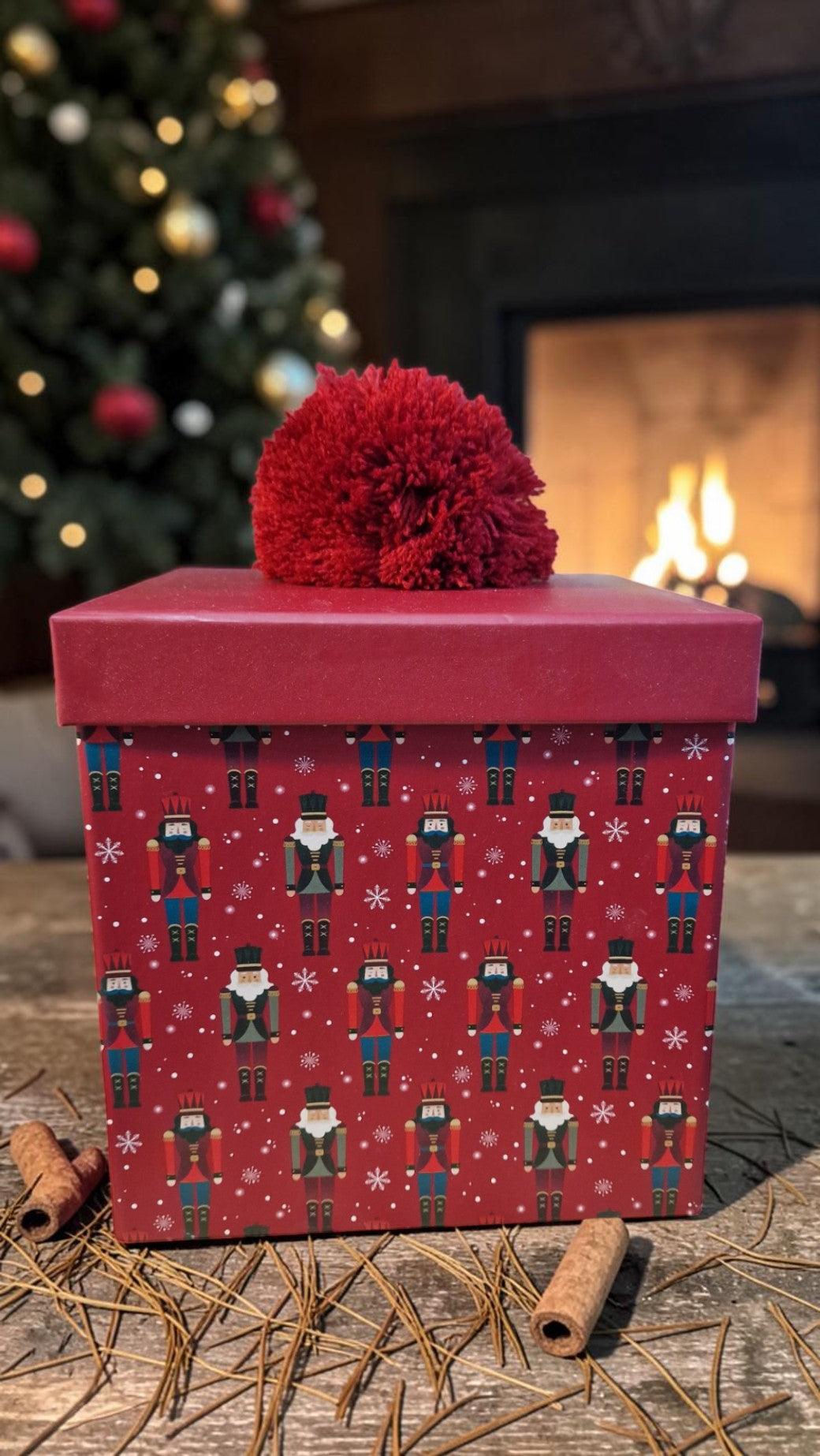 Red gift box with nutcracker design on a wooden surface with a Christmas tree and fireplace in the background.