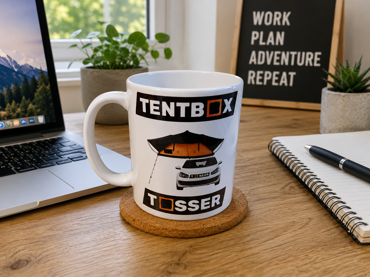A white ceramic mug with a tentbox car graphic and the text 'TENTBOX' and 'TOSSER' sits on a cork coaster on a wooden desk. The scene is styled with a laptop, notebook, pen, and potted plants in a bright home office environment.